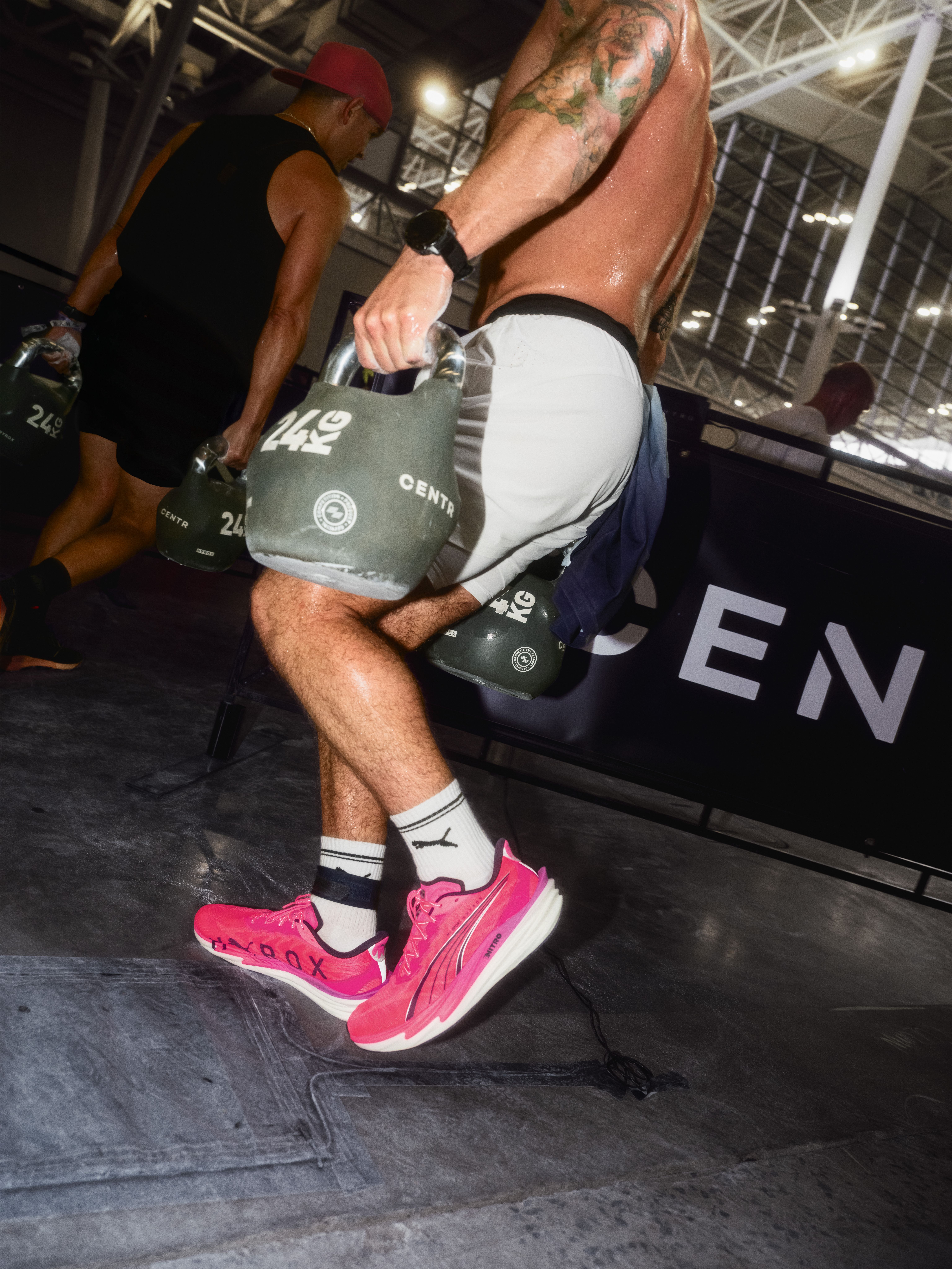 Two sweaty men, one in bright pink Puma shoes, carry heavy 24kg kettlebells across a gym floor.