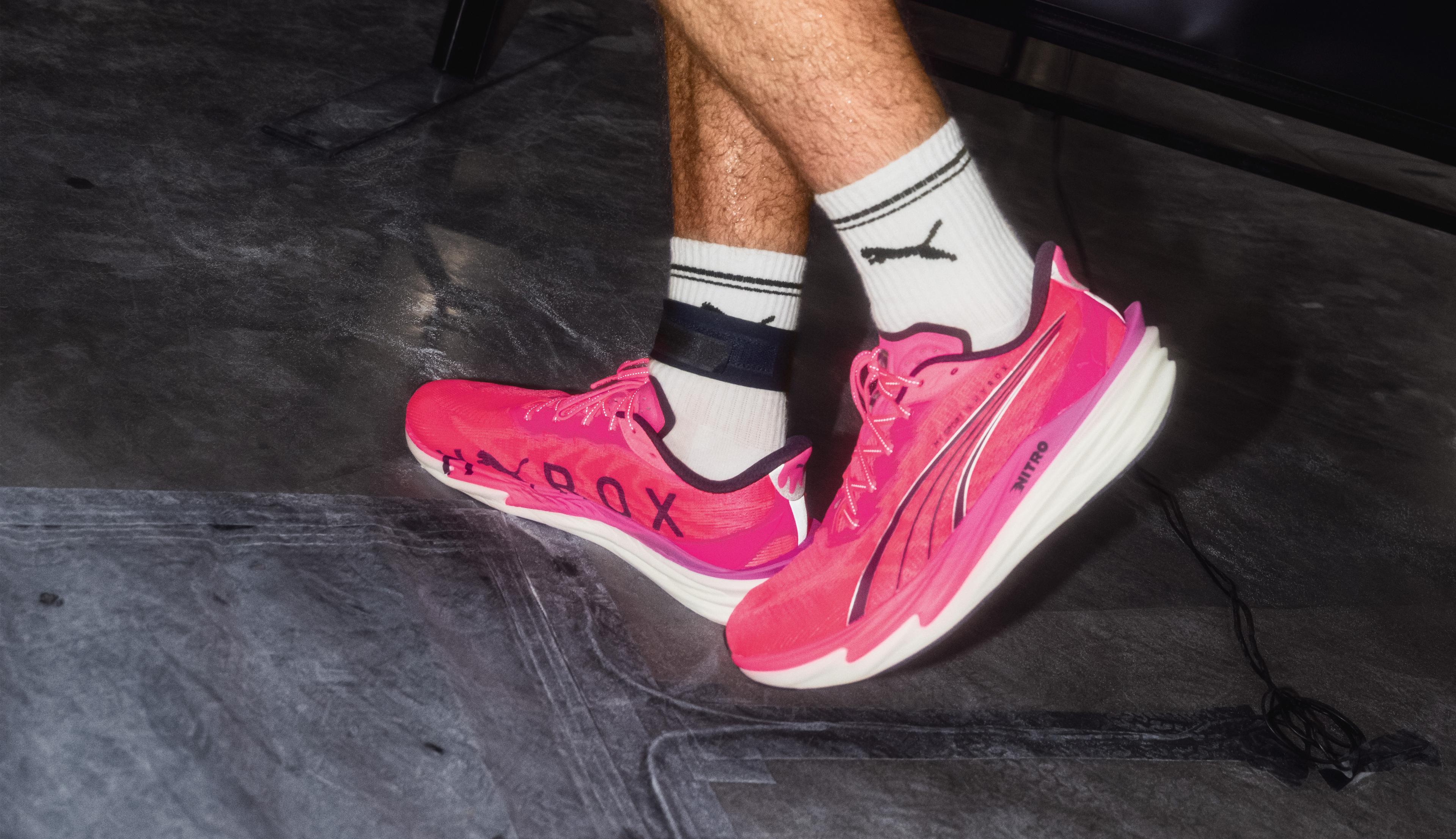 Sweaty athlete in pink sneakers carries two heavy kettlebells indoors.