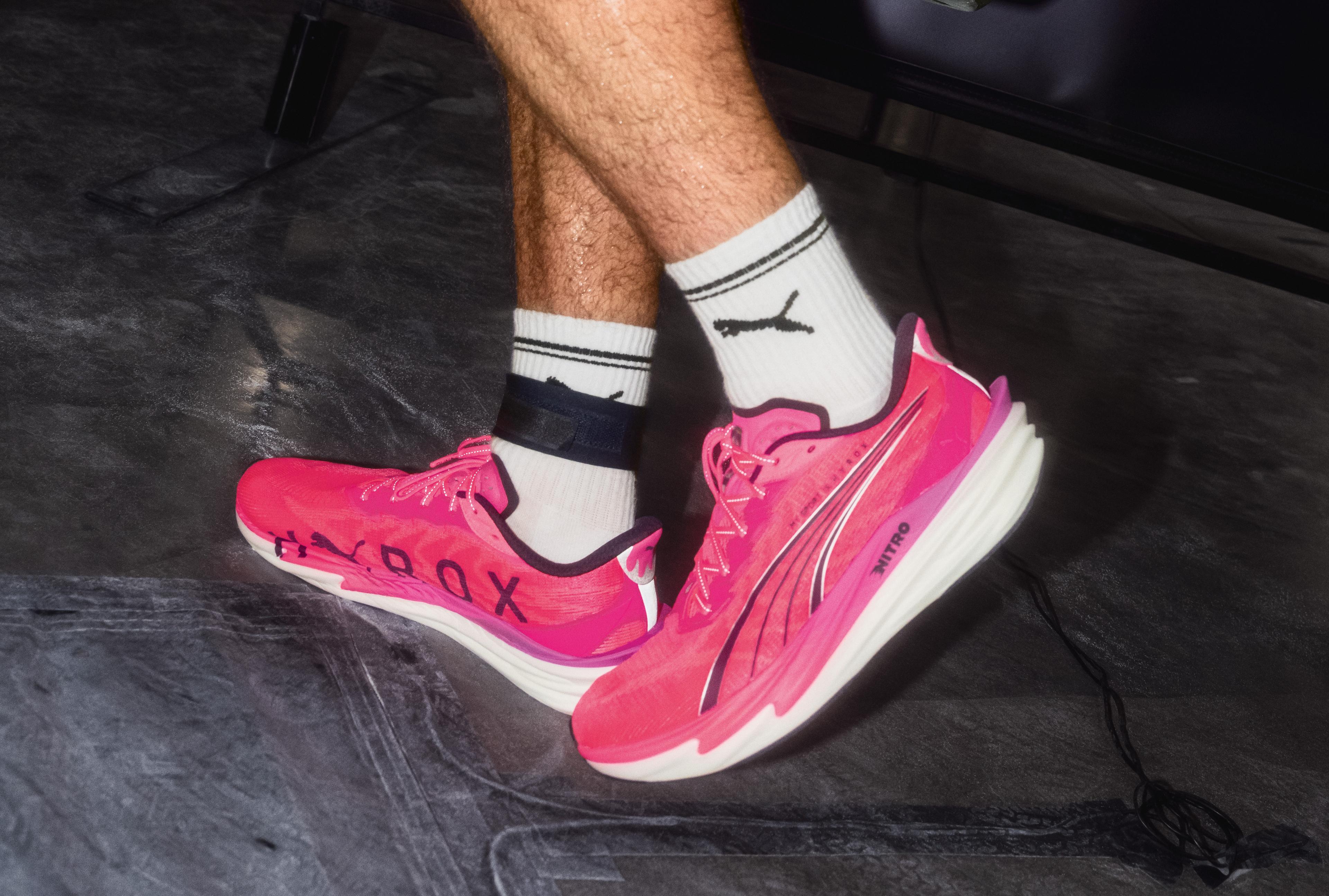 Two sweaty men, one in bright pink Puma shoes, carry heavy 24kg kettlebells across a gym floor.
