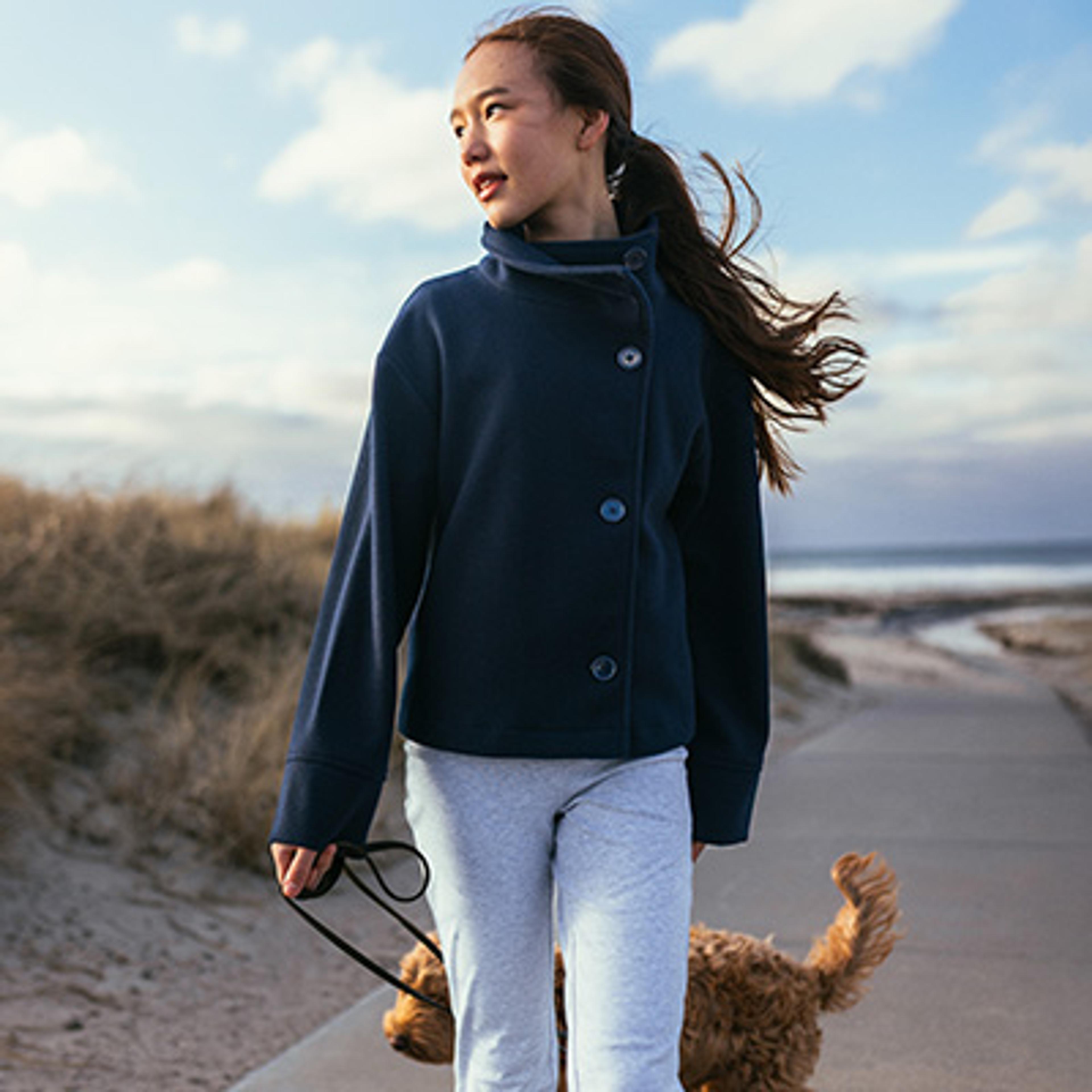 A young girl in a blue jacket walks her fluffy brown dog on a beachside path.