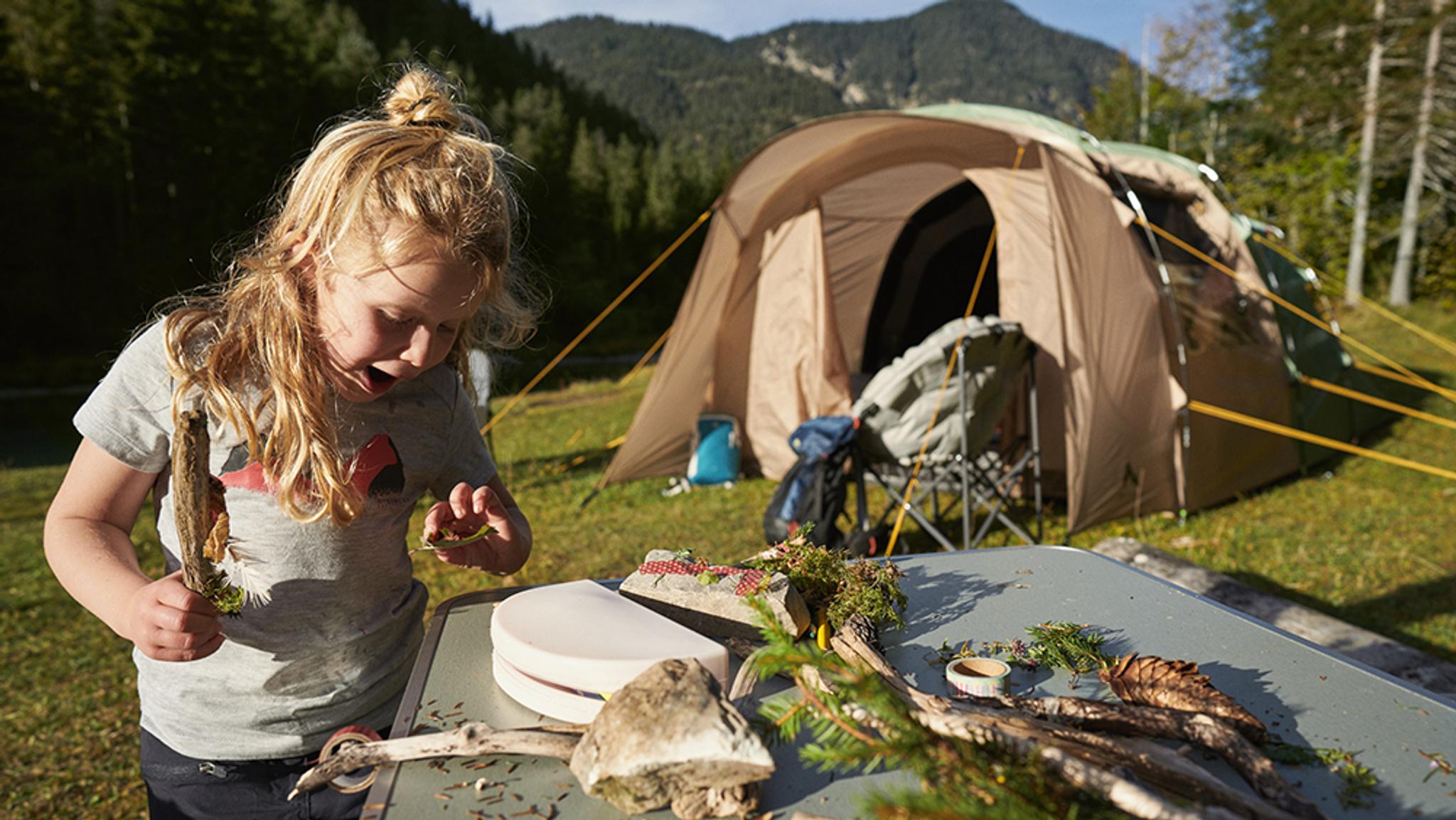 A blonde child intently building with sticks and pinecones on a table at a sunny mountain campsite.