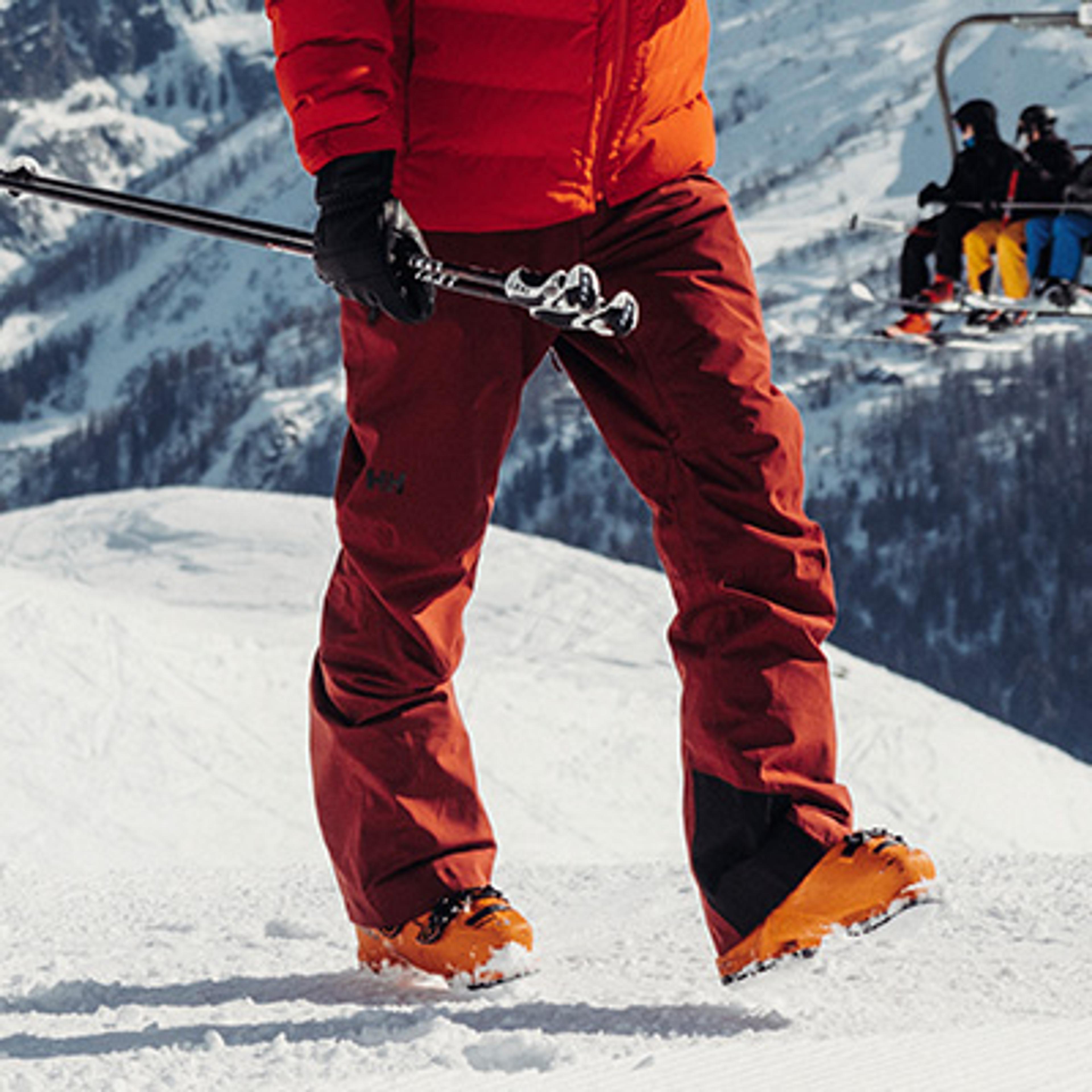 A skier in a red jacket and brown pants holds ski poles on a snowy slope with a ski lift behind them.
