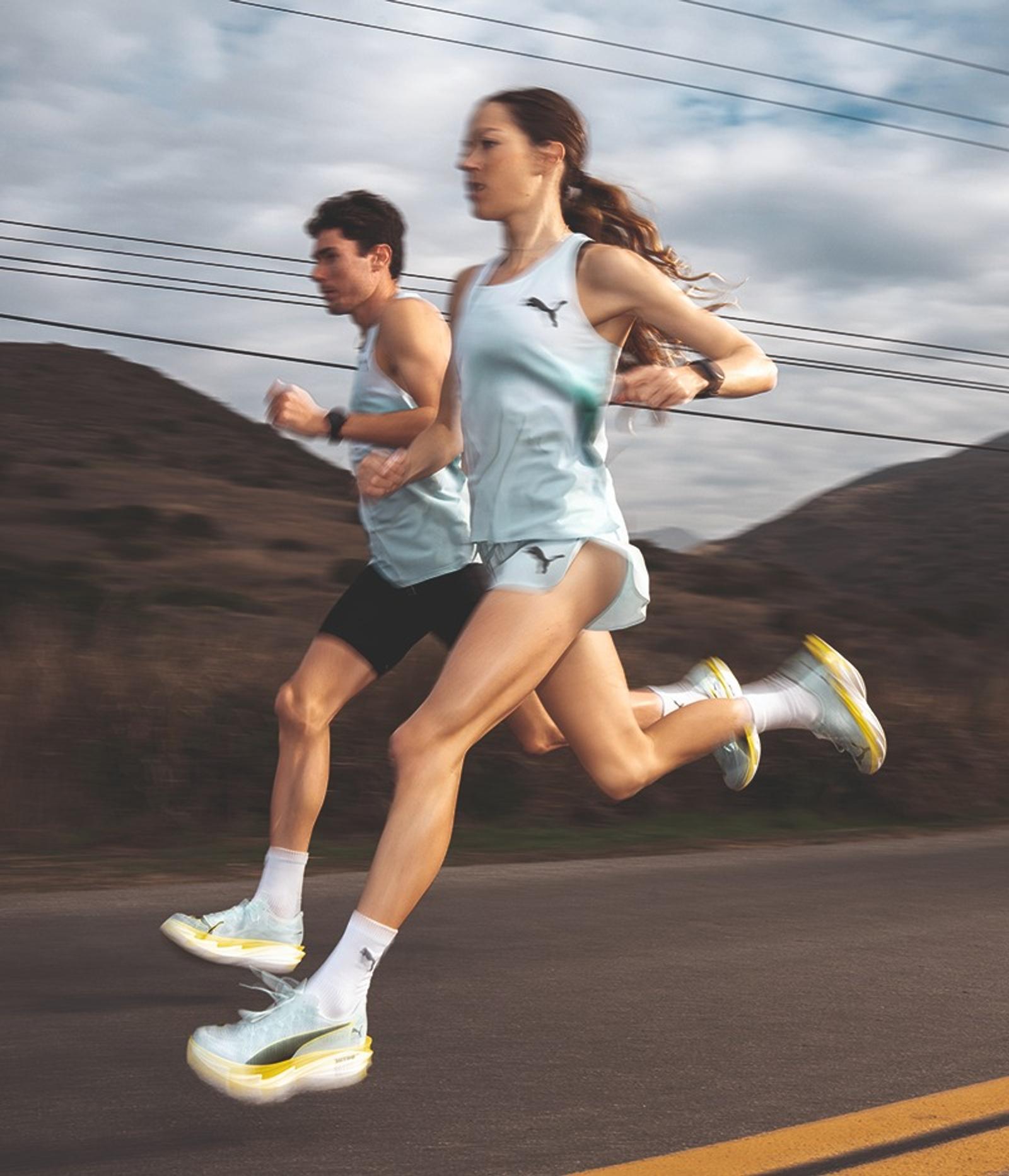 Two runners, a woman and a man, in light blue Puma athletic wear and shoes, running on a road with blurred hills and power lines in the background.