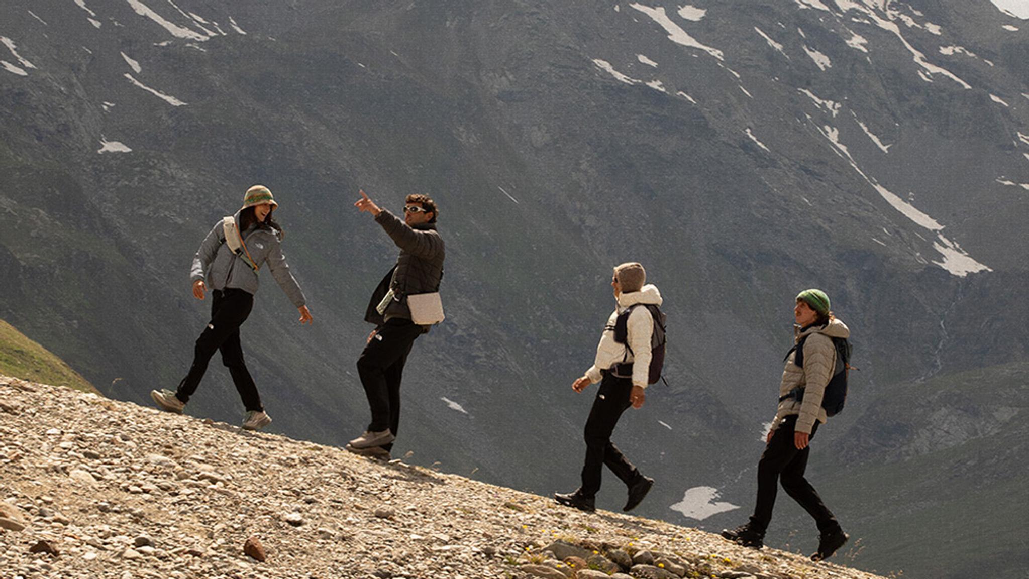 Four hikers ascend a rocky mountain slope, with one person pointing towards a distant snowy peak.