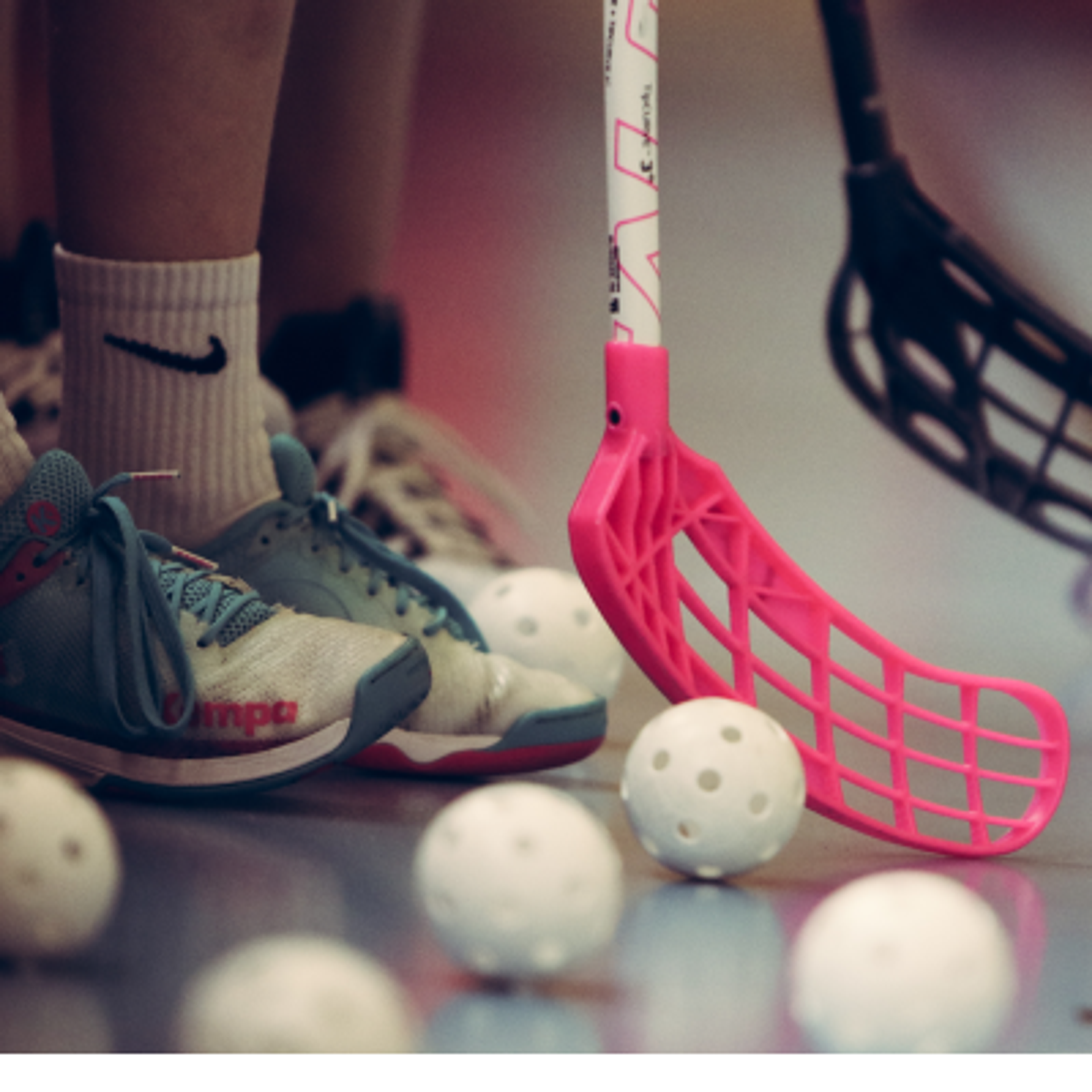 Player's feet in athletic shoes next to a pink floorball stick and several white floorball balls.