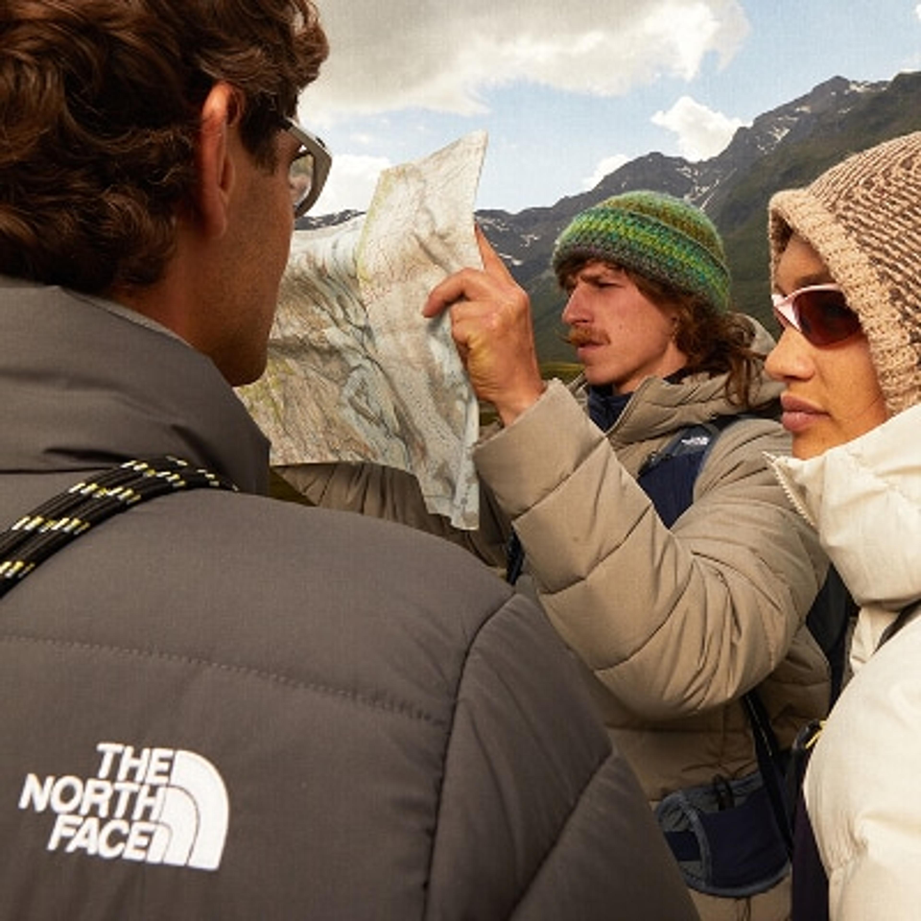 Three people wearing winter gear study a map against a mountain backdrop. A North Face logo is visible.