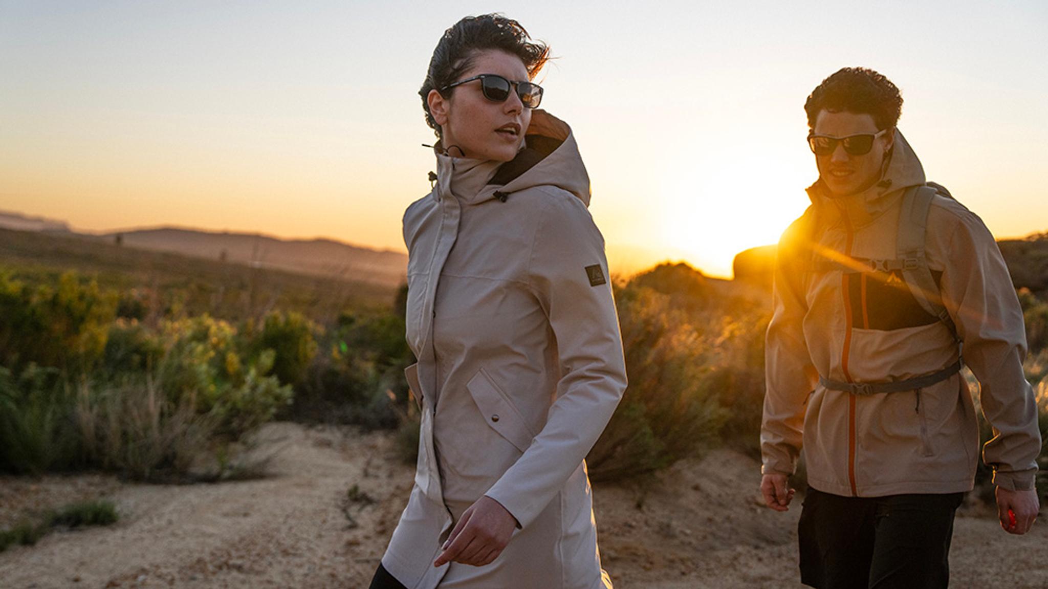 Two hikers in jackets and sunglasses walk a dirt path at sunset.