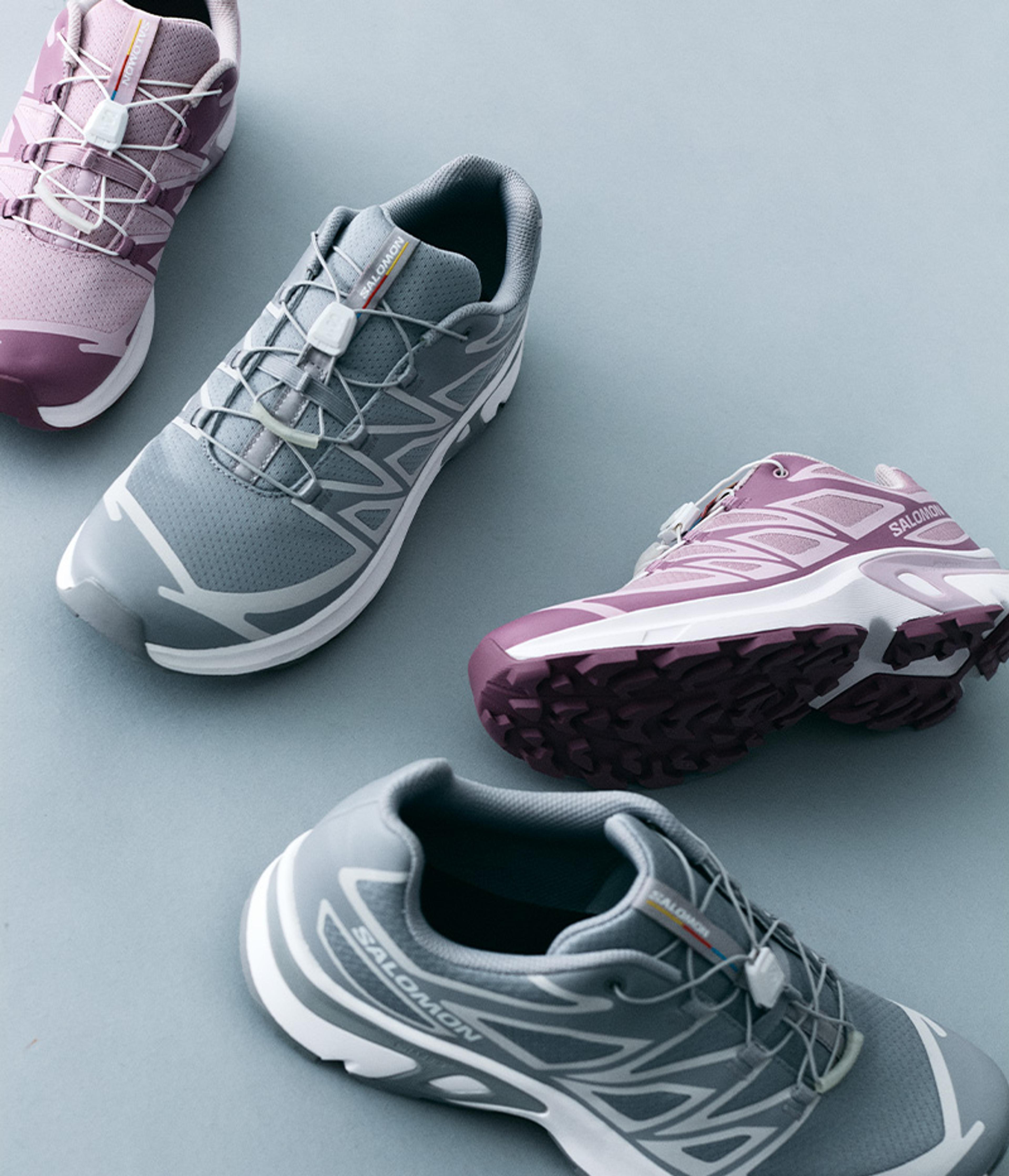 Four Salomon trail running shoes, two grey and two purple, with quick-lace systems, on a light background.