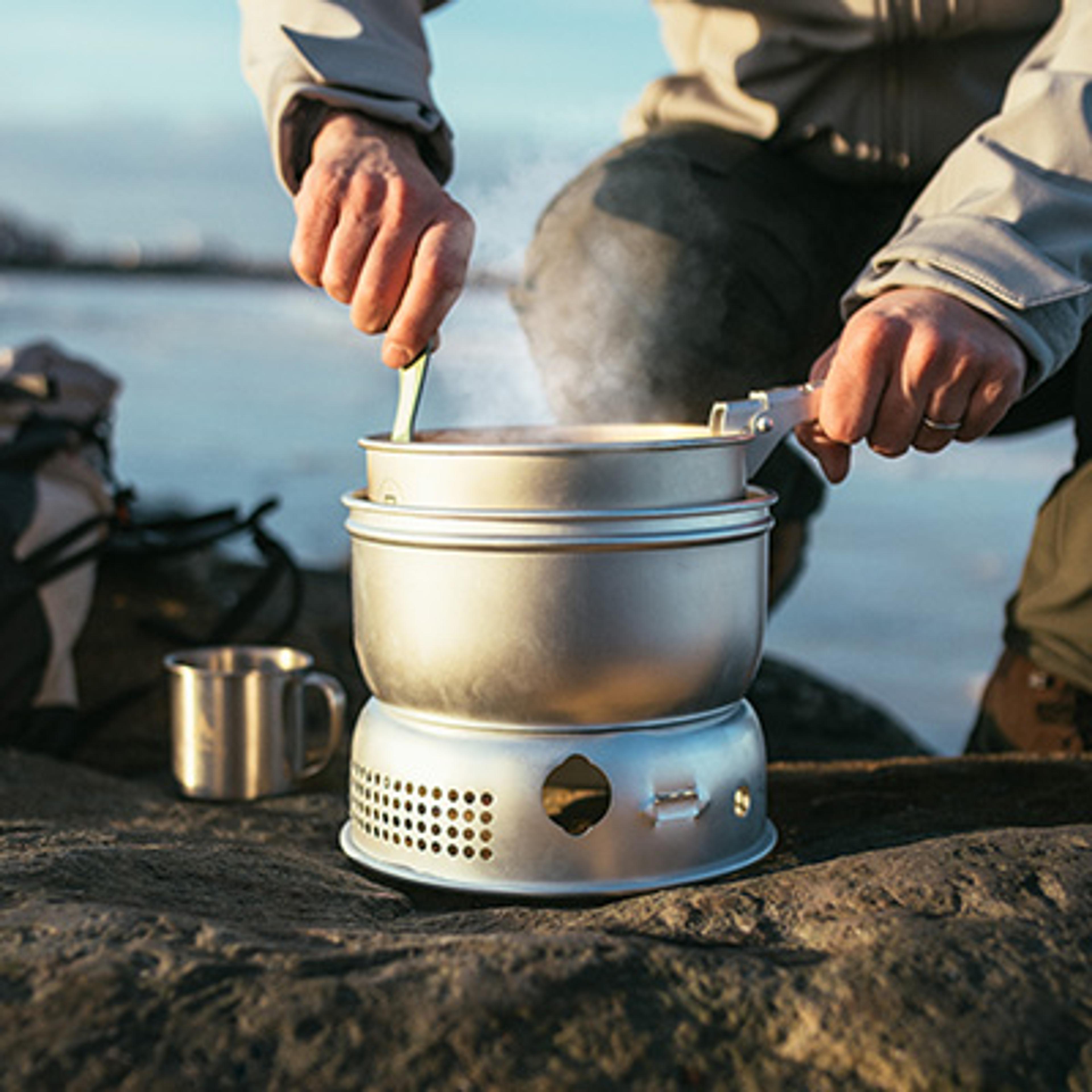 Hands stir a steaming pot on an outdoor camping stove.