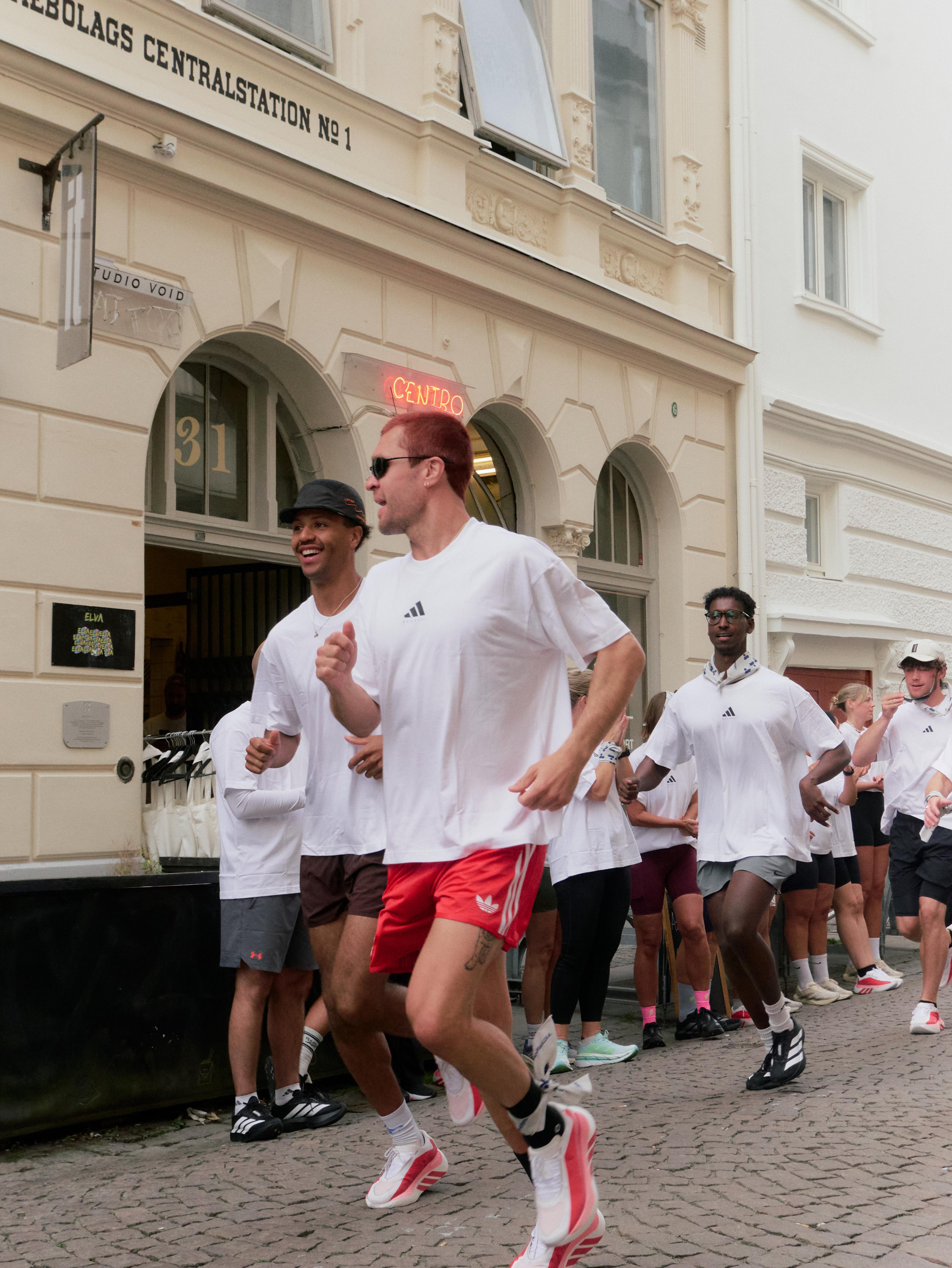 A group of diverse runners in white Adidas shirts and athletic shorts runs down a cobblestone street past a building.