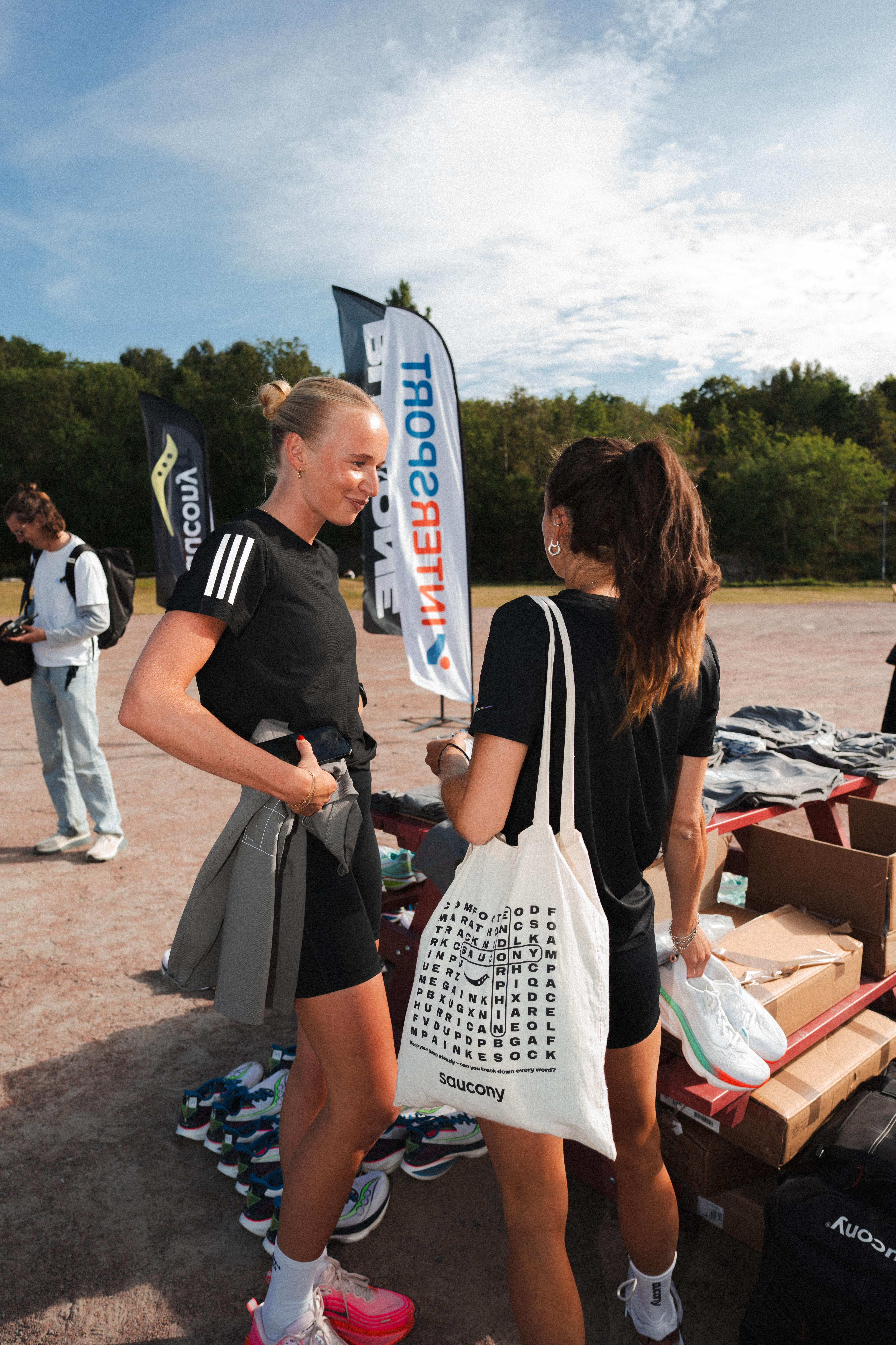 Two women in athletic wear chat at an outdoor event surrounded by running shoes and brand flags, including Saucony and Intersport.
