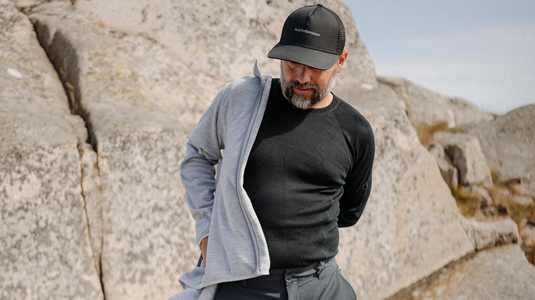 A man in a black shirt and cap putting on a light grey jacket outdoors amidst large rocks.