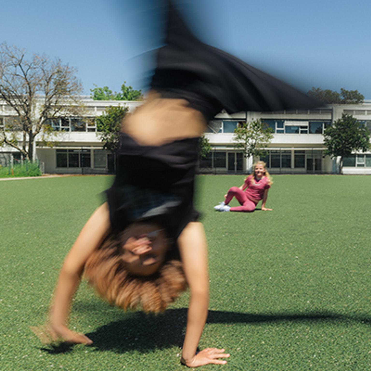 a girl is doing a handstand in a field while another girl sits on the grass .