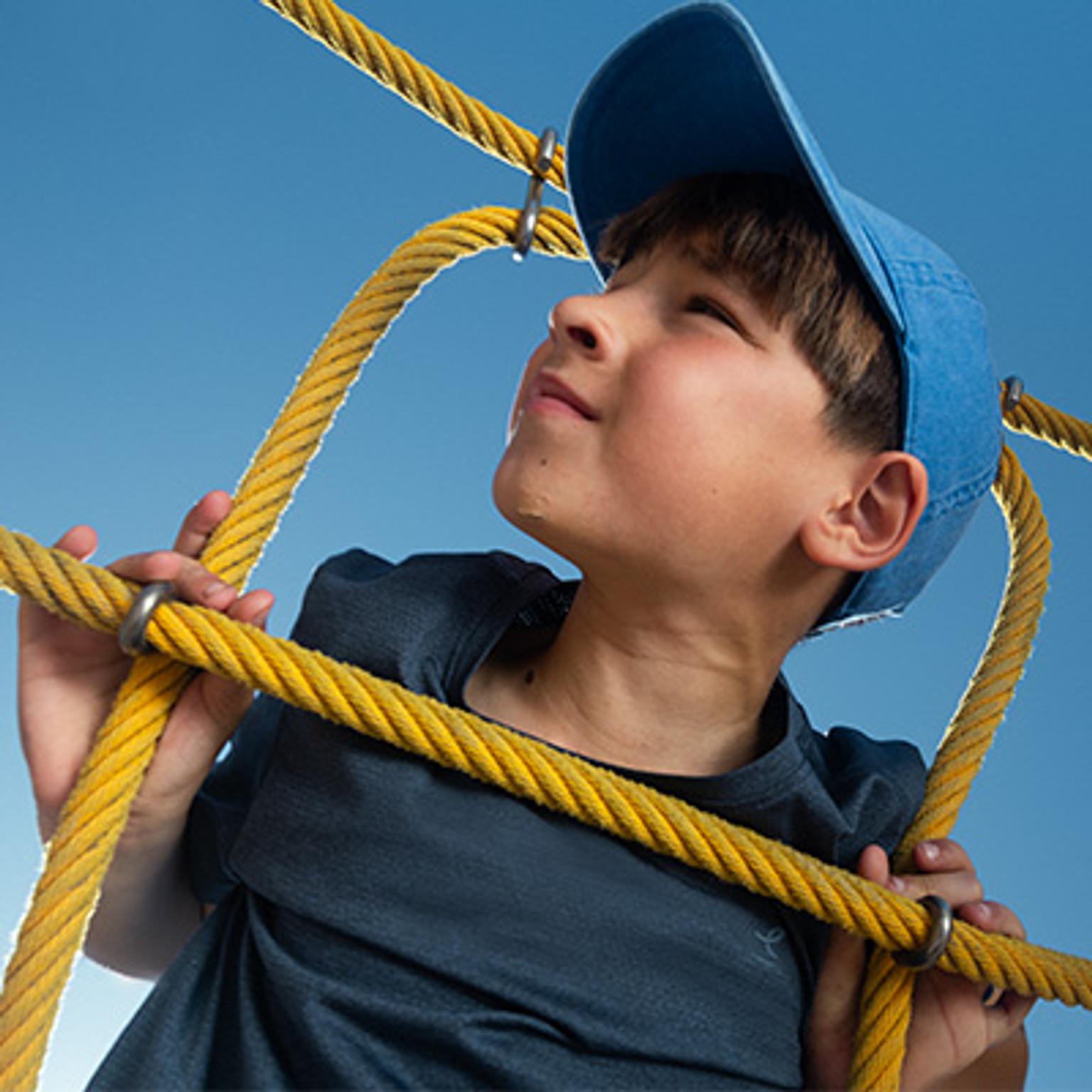 A young boy in a blue cap holds yellow ropes, looking up at a clear blue sky.
