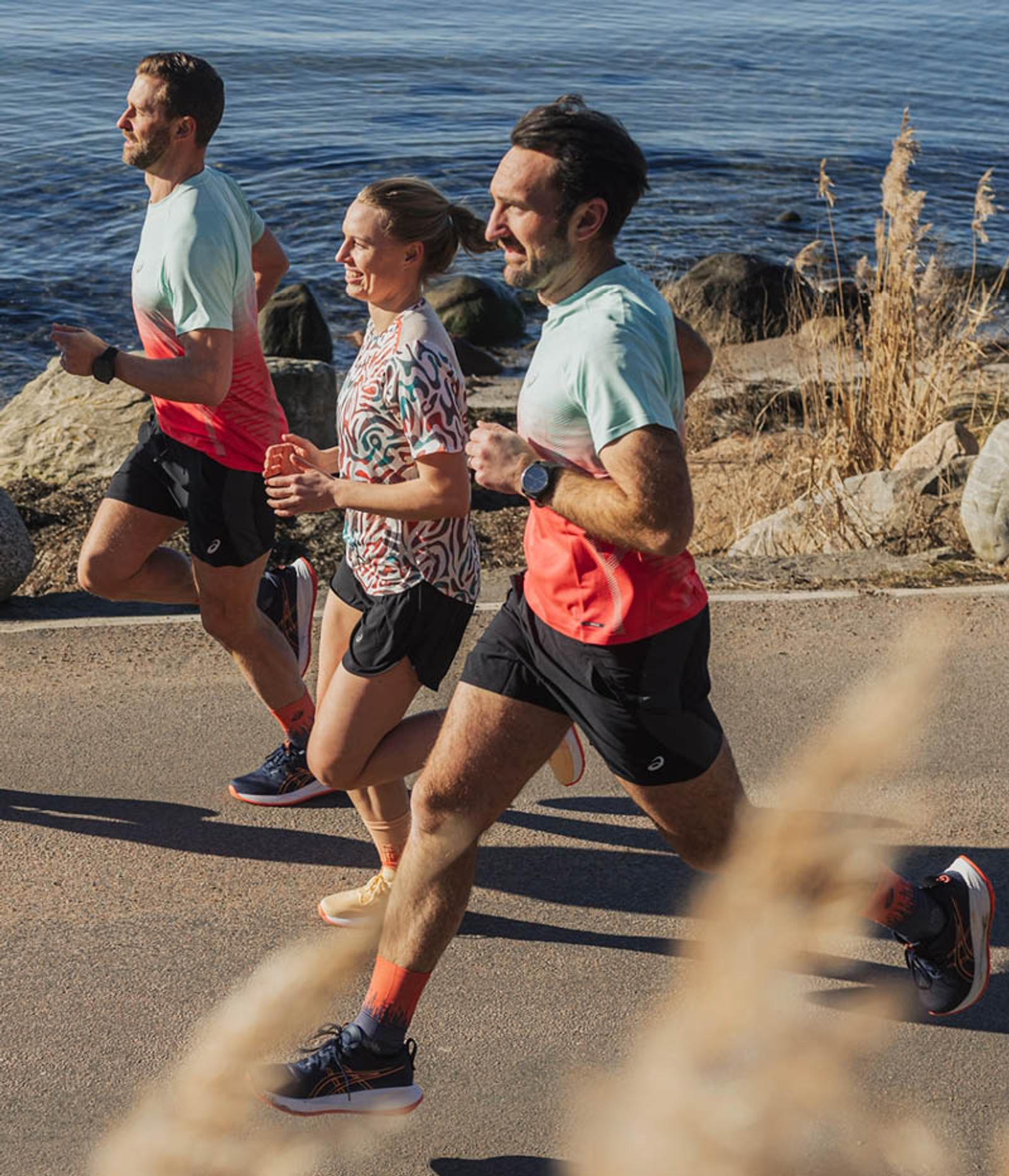 Three smiling runners on a paved path by the water on a sunny day.