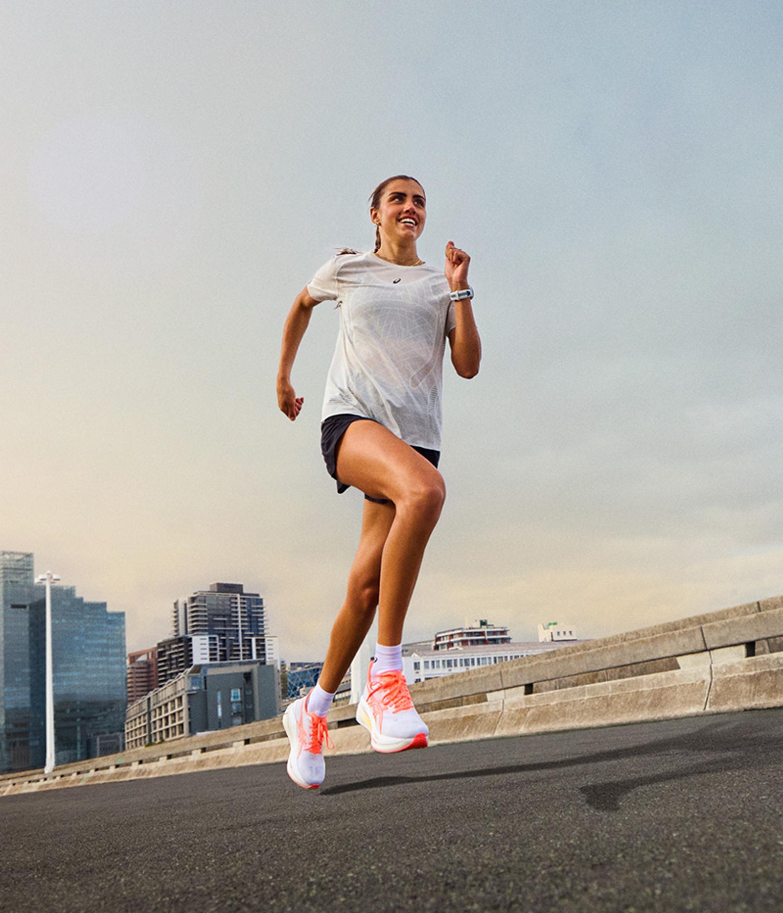A smiling woman in a white t-shirt and black shorts runs on a city road with buildings in the background.
