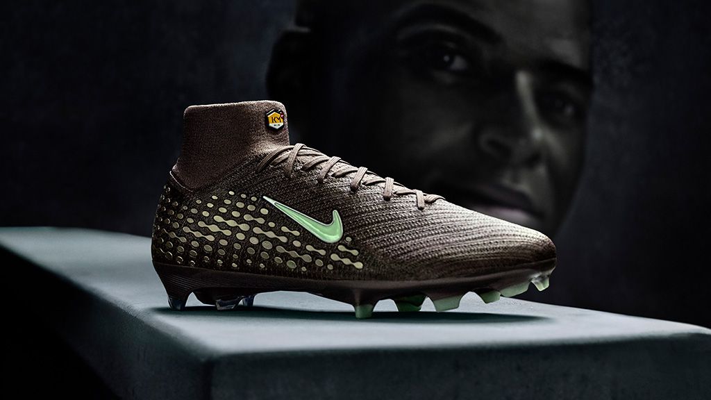 Dark brown Nike soccer cleat with light green swoosh, dotted pattern, and studs, featuring a high collar with a TN logo, against a dark silhouetted face.