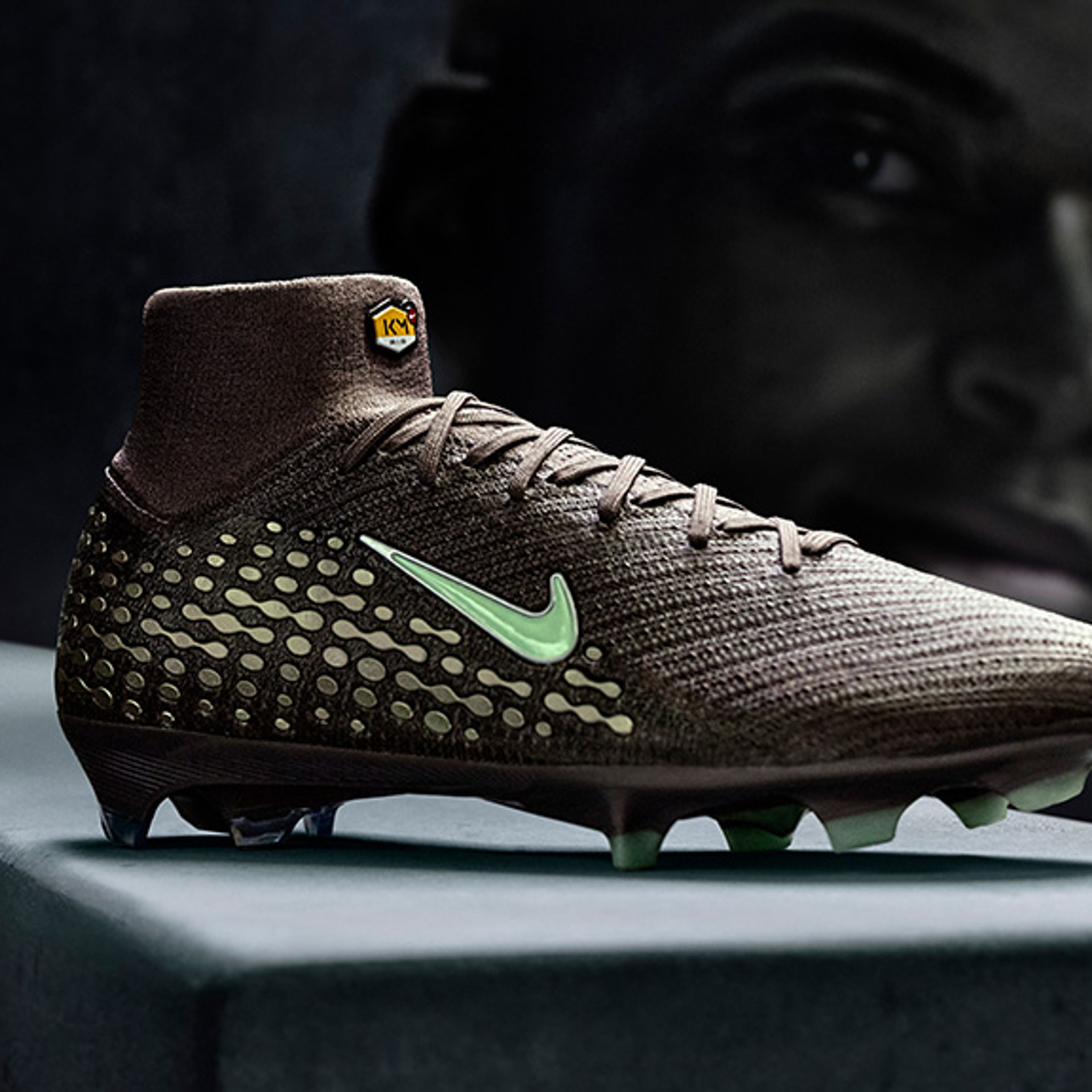 Dark brown Nike soccer cleat with light green swoosh, dotted pattern, and studs, featuring a high collar with a TN logo, against a dark silhouetted face.