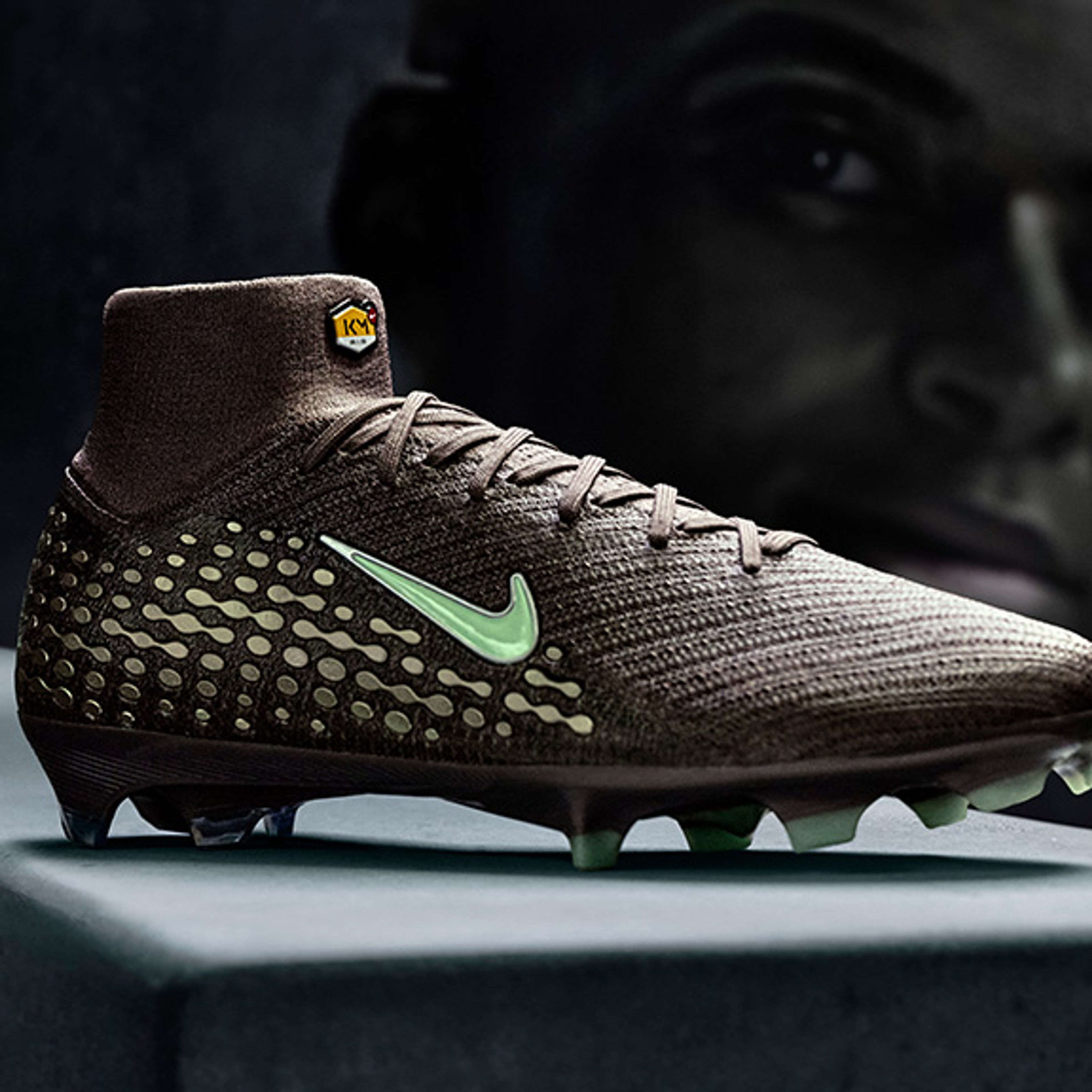 Dark brown football boot with a light green Nike swoosh, studded sole, and ankle collar, in front of a dark, blurry face.