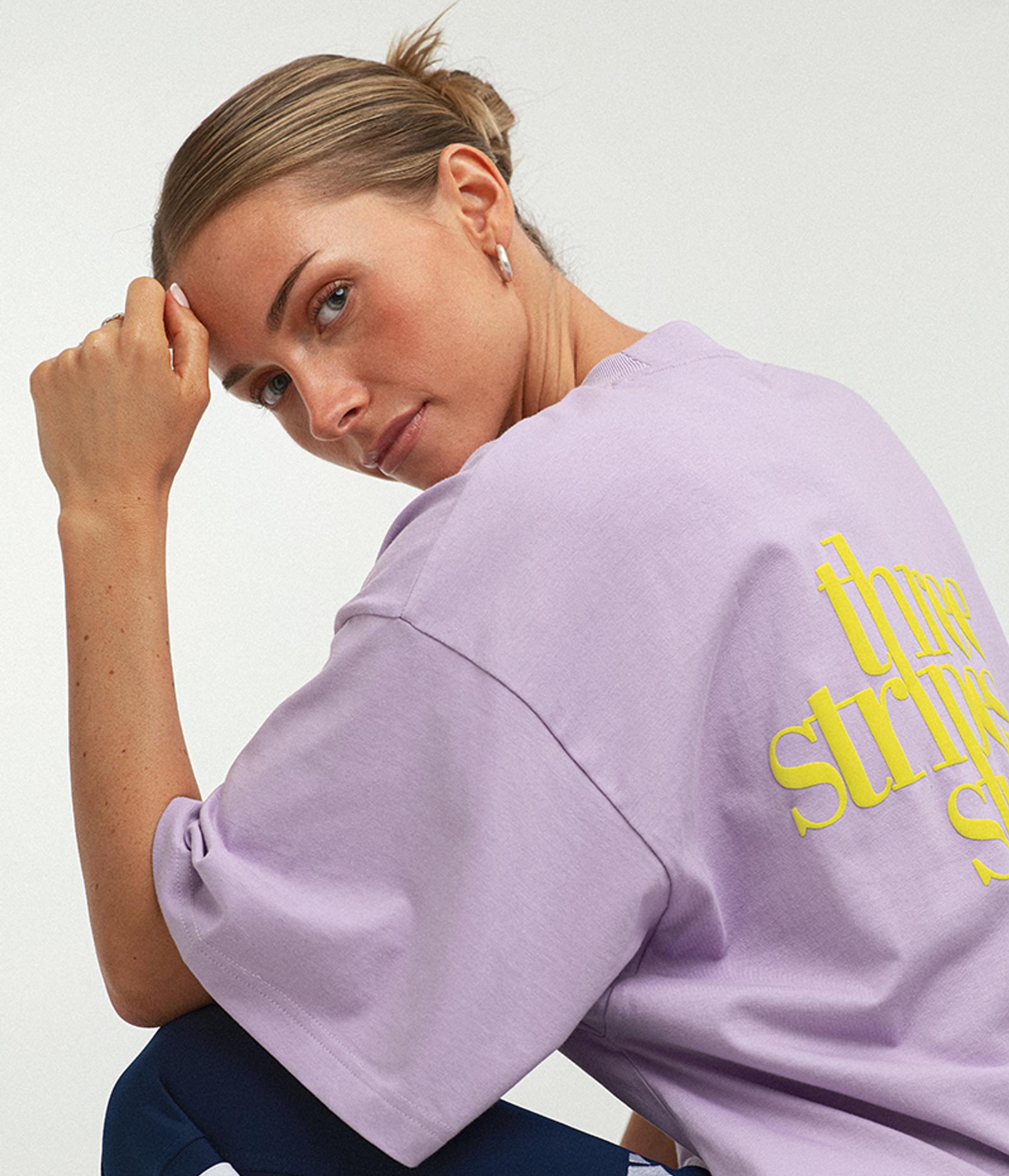 A woman in a light purple t-shirt with yellow "three stripes" text on the back looks over her shoulder.