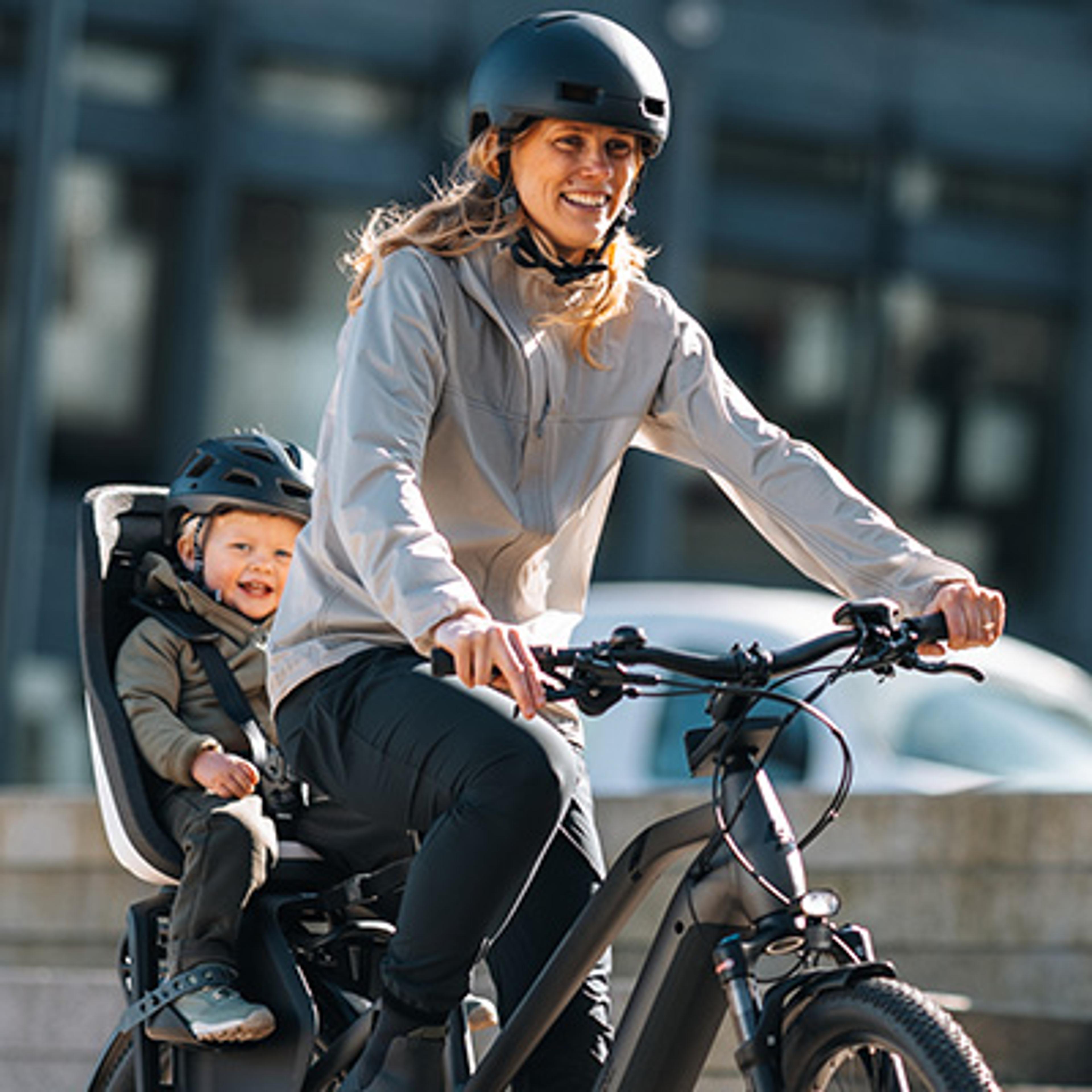 A woman on a bicycle with a child in a rear seat, both wearing helmets and smiling happily.