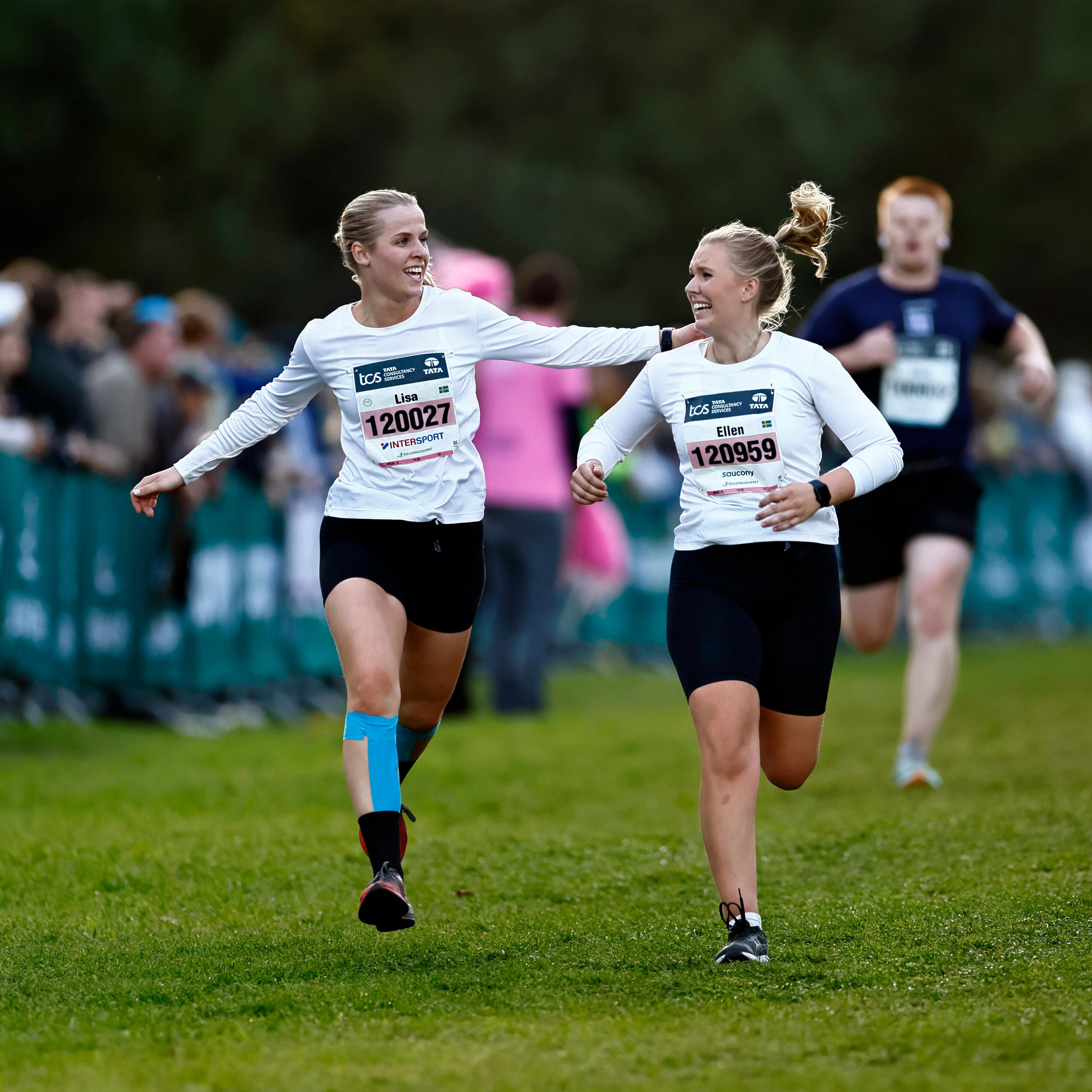 Two smiling women run a race together on a grassy field.