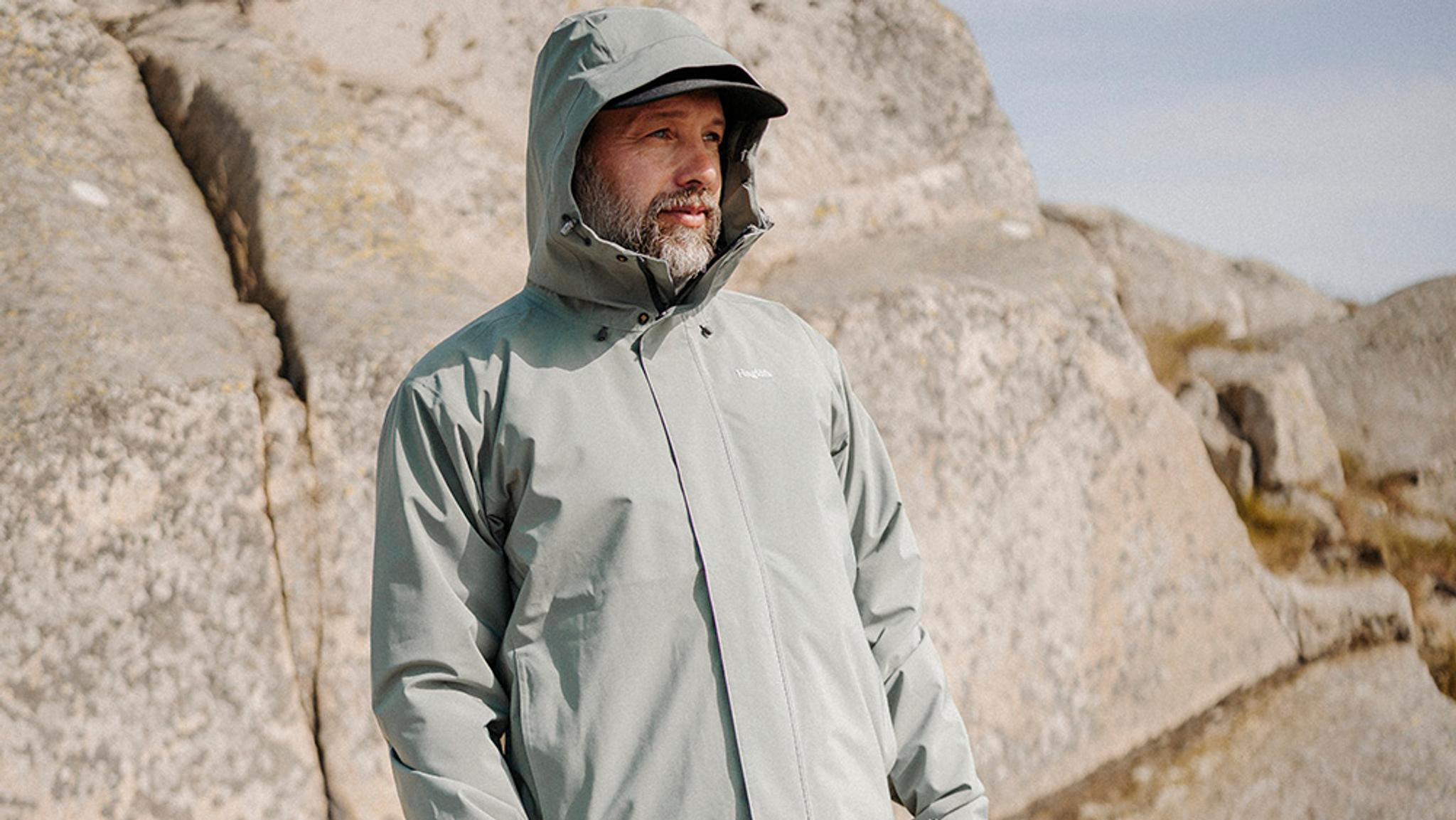 A bearded man in a grey hooded jacket and cap stands outdoors against a backdrop of rocks.