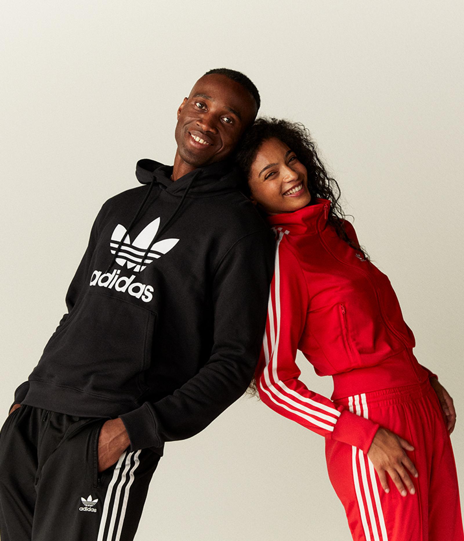 a man wearing a black adidas hoodie stands next to a woman wearing red adidas pants