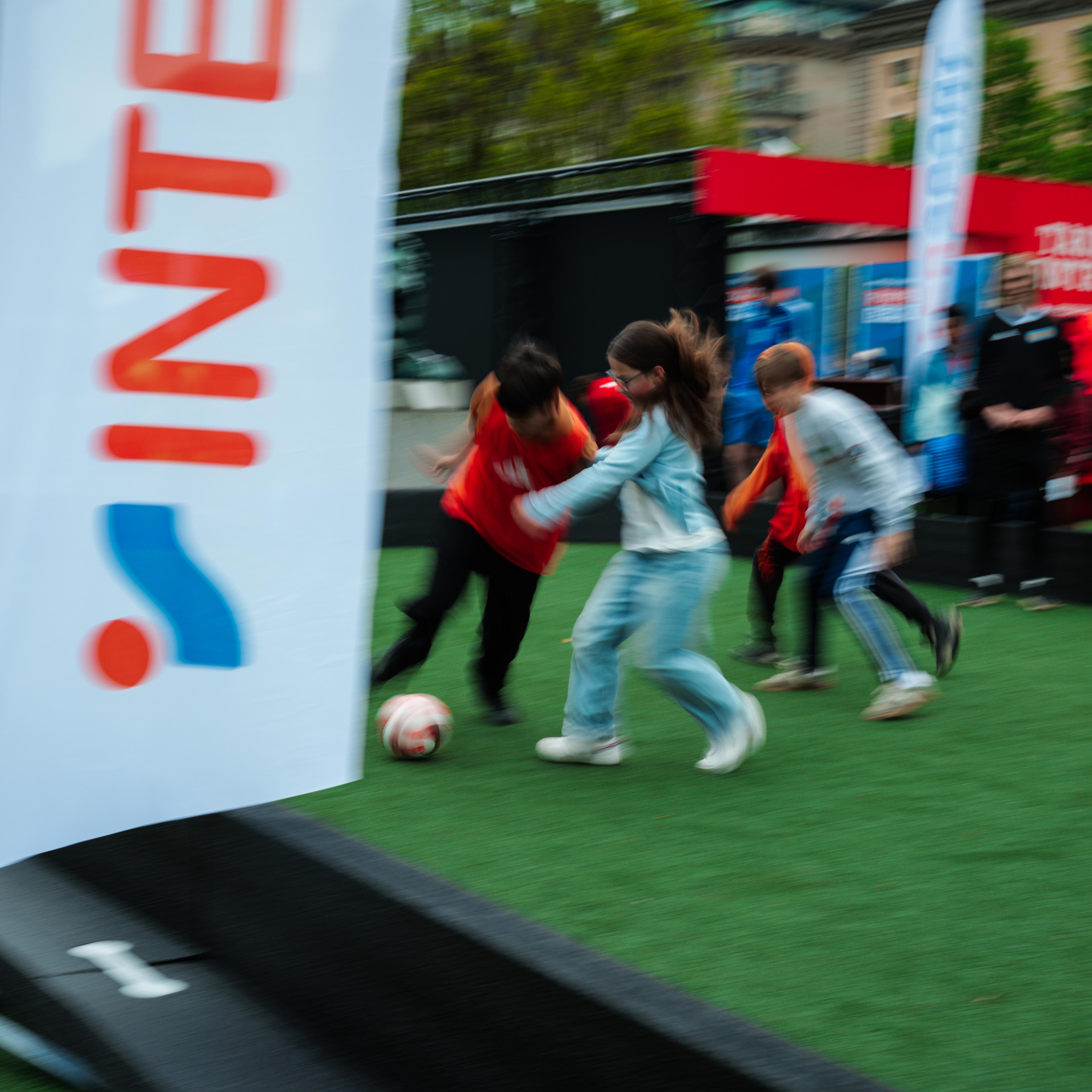 Children playing soccer on a green turf with motion blur, an Intersport banner visible.
