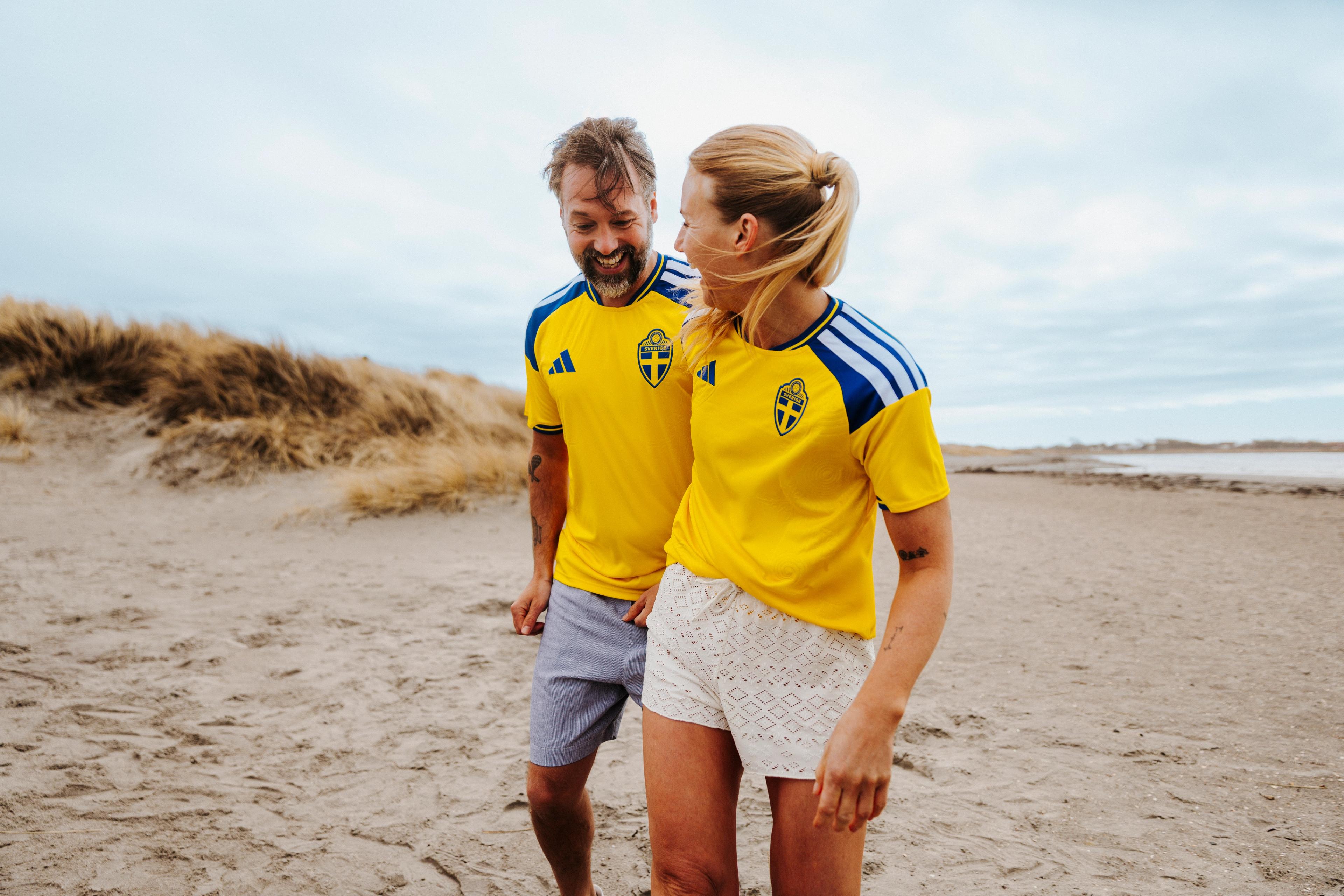 A smiling man and woman wearing yellow Swedish national team jerseys walk on a sandy beach with dunes.