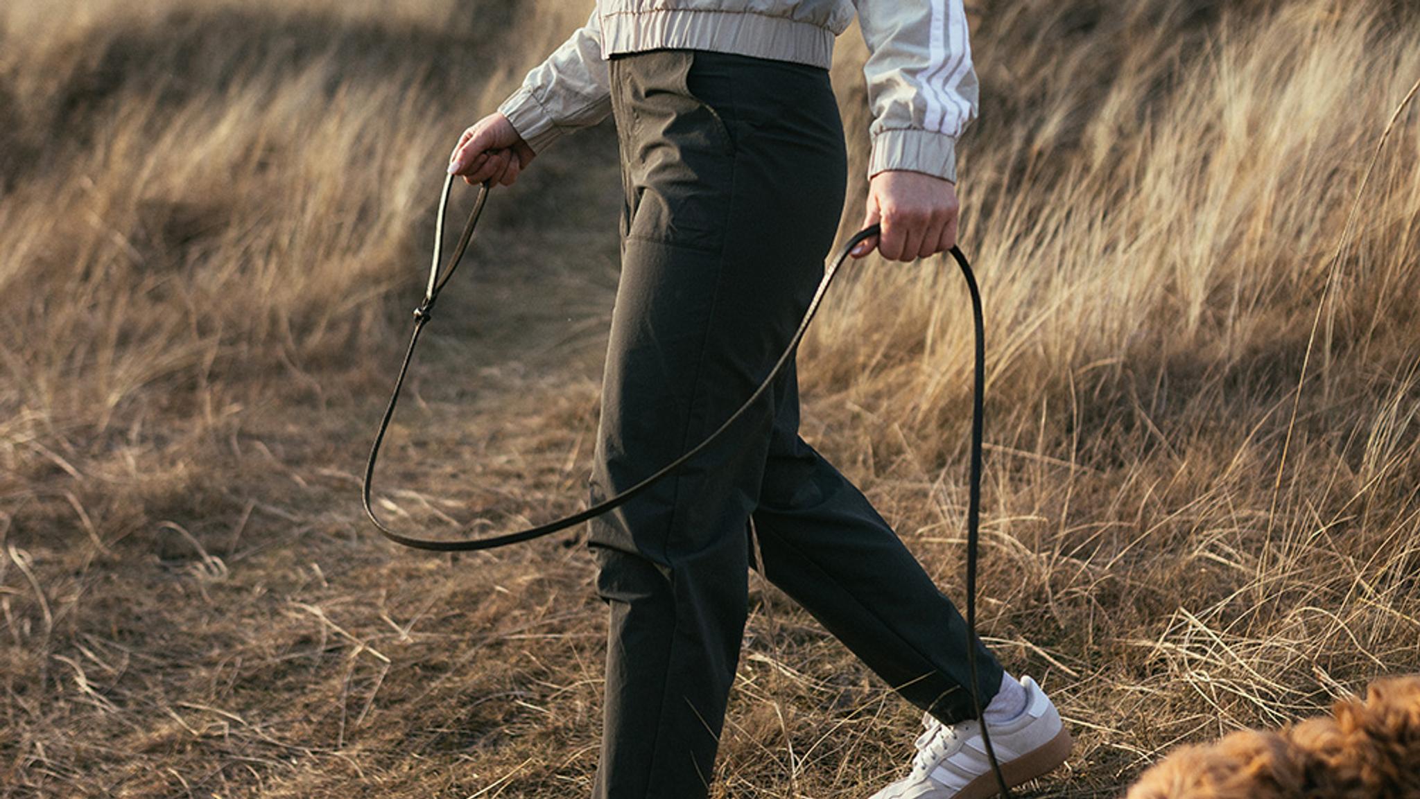 Person walking a dog on a leash through dry grass with her green outdoor pants