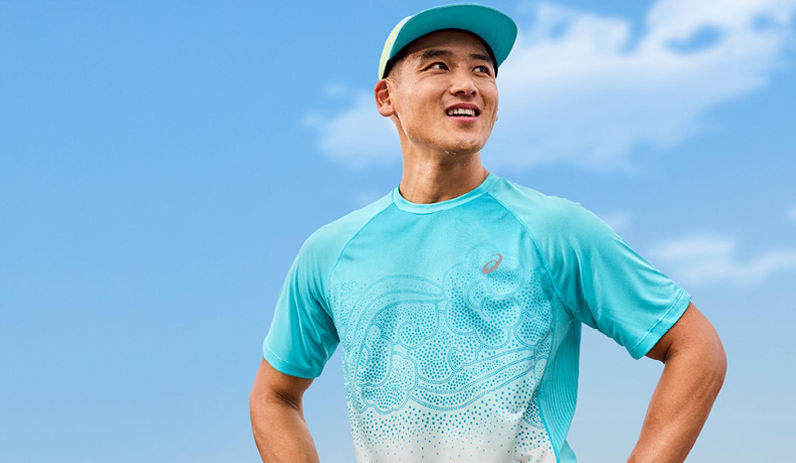 A smiling man in a turquoise ASICS running shirt and cap against a blue sky.