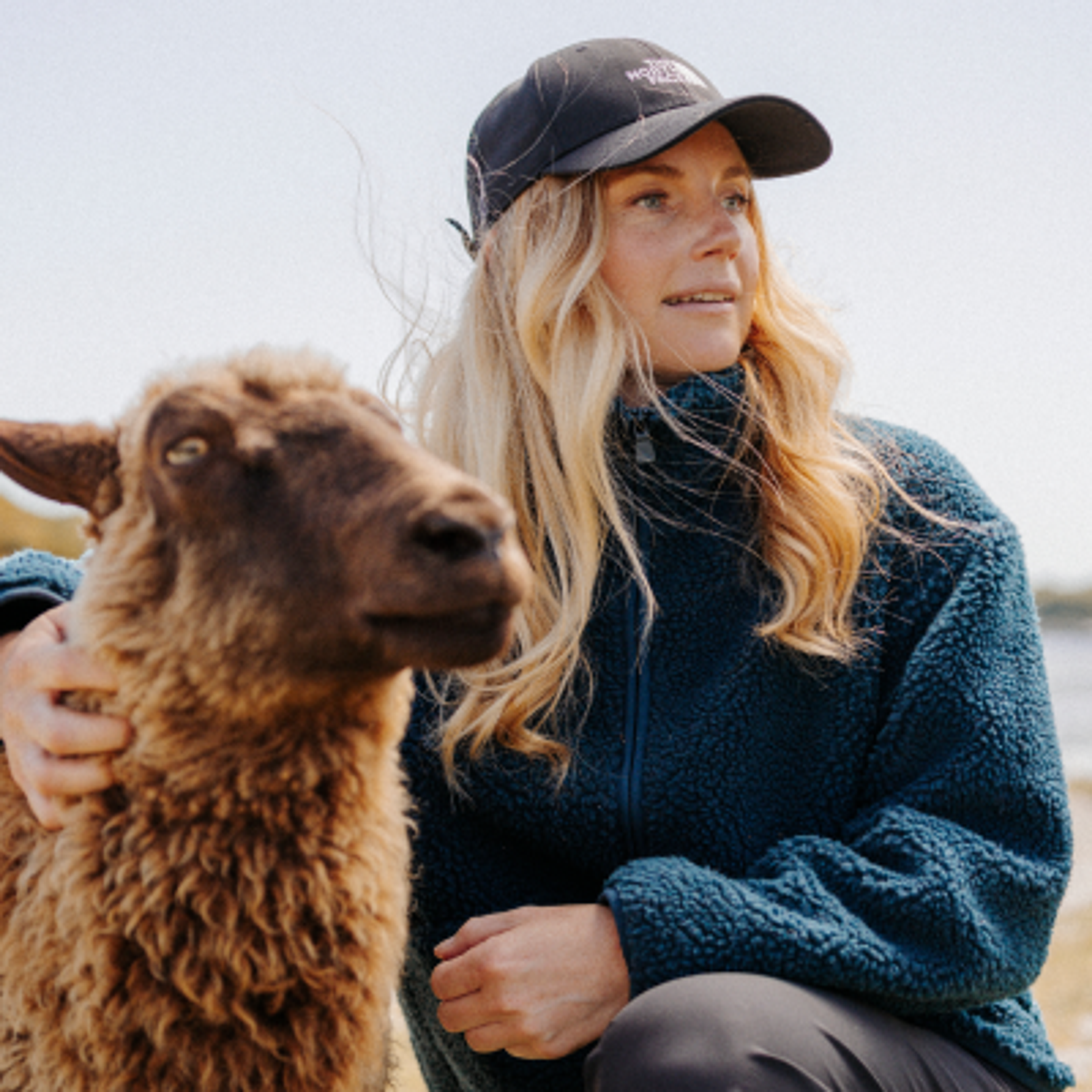 A woman in a cap and blue fleece jacket pets a brown lamb outdoors.