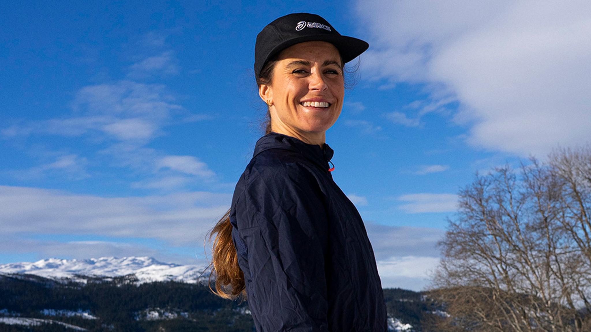 Woman, Anna Haag, in a cap and jacket with snowy mountains and a blue sky in the background.