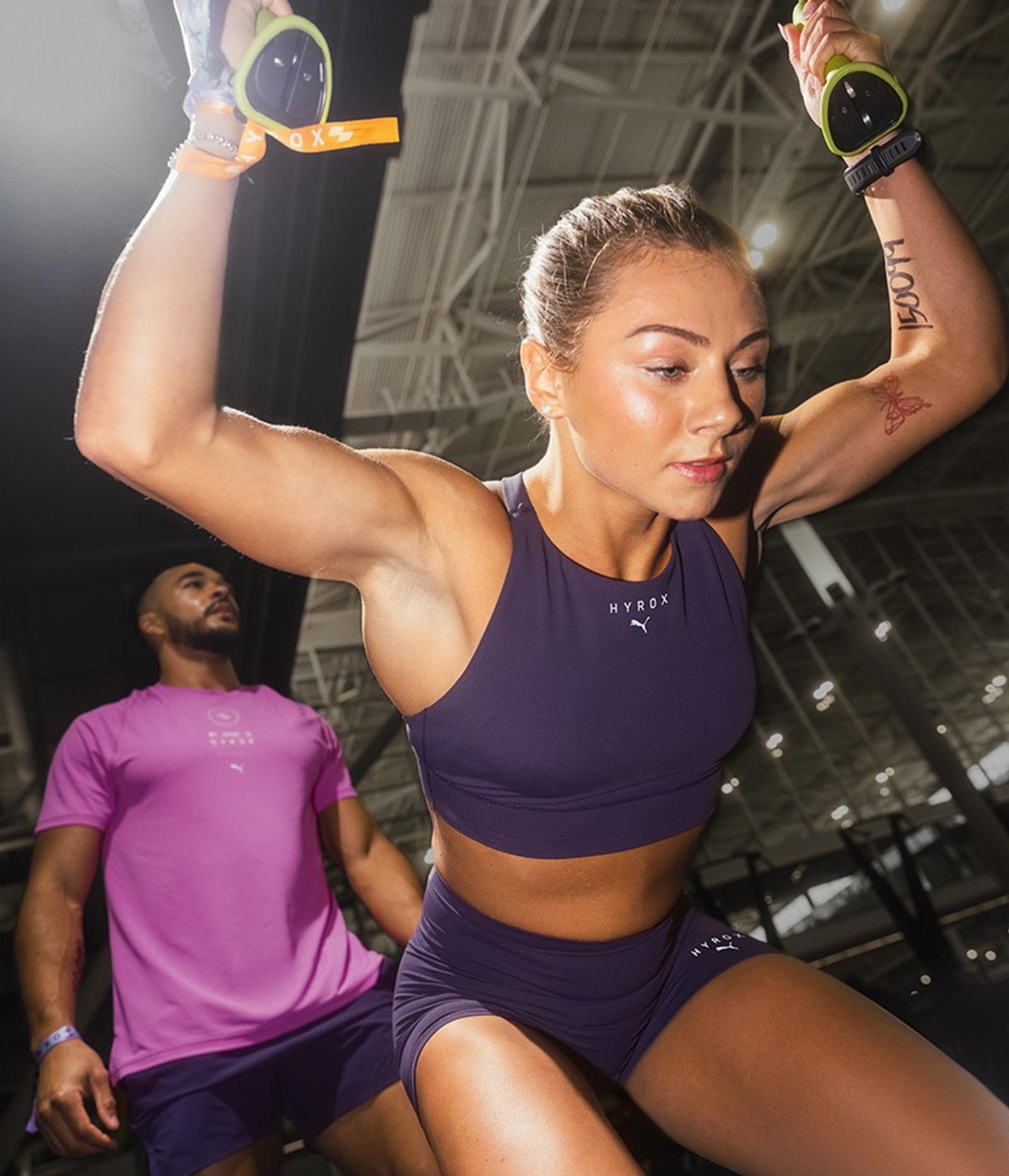 A focused woman in a purple sports bra and shorts pulls down on exercise equipment, with a man blurred in the background.