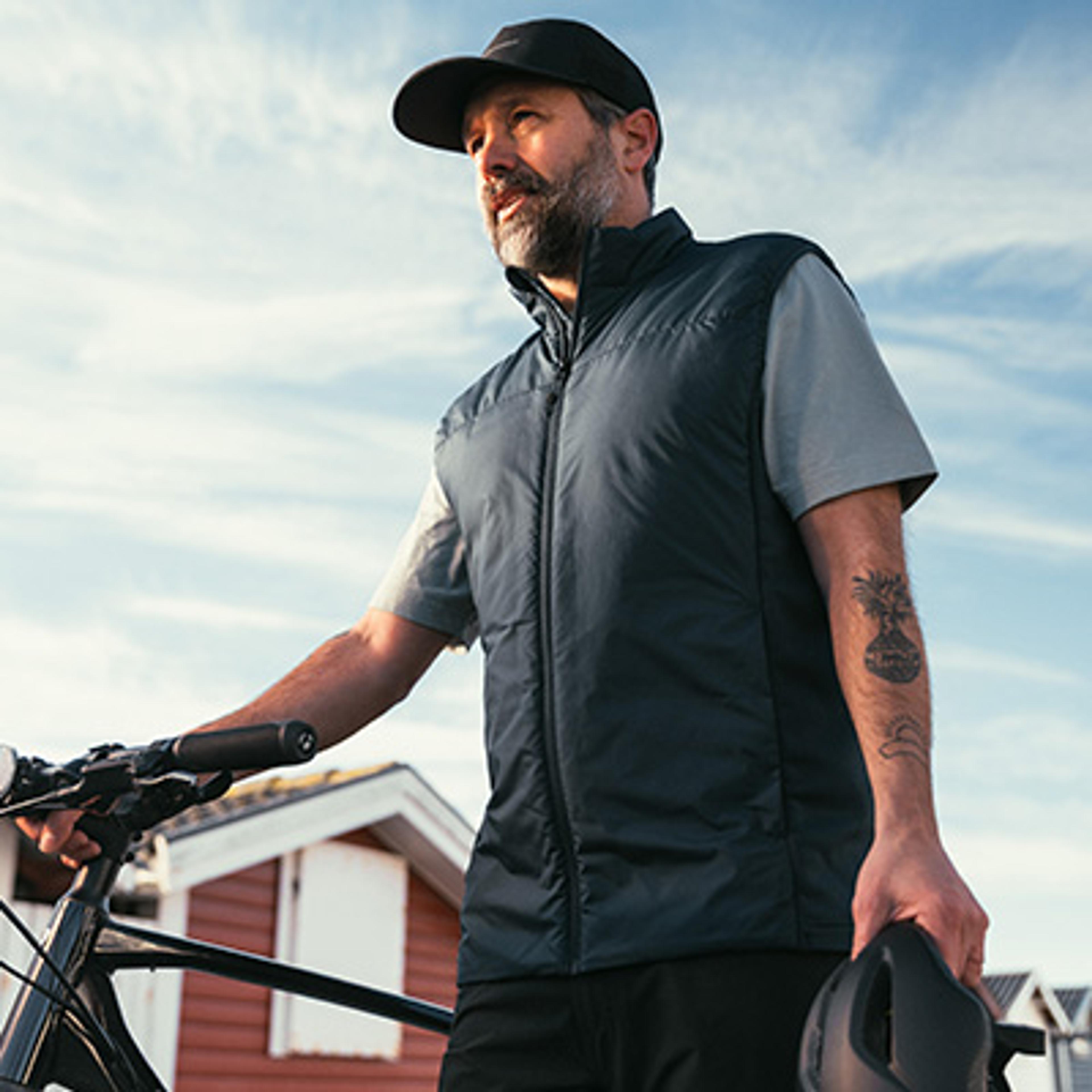 Bearded man in a cap and dark vest holding a bike and helmet, looking upwards.