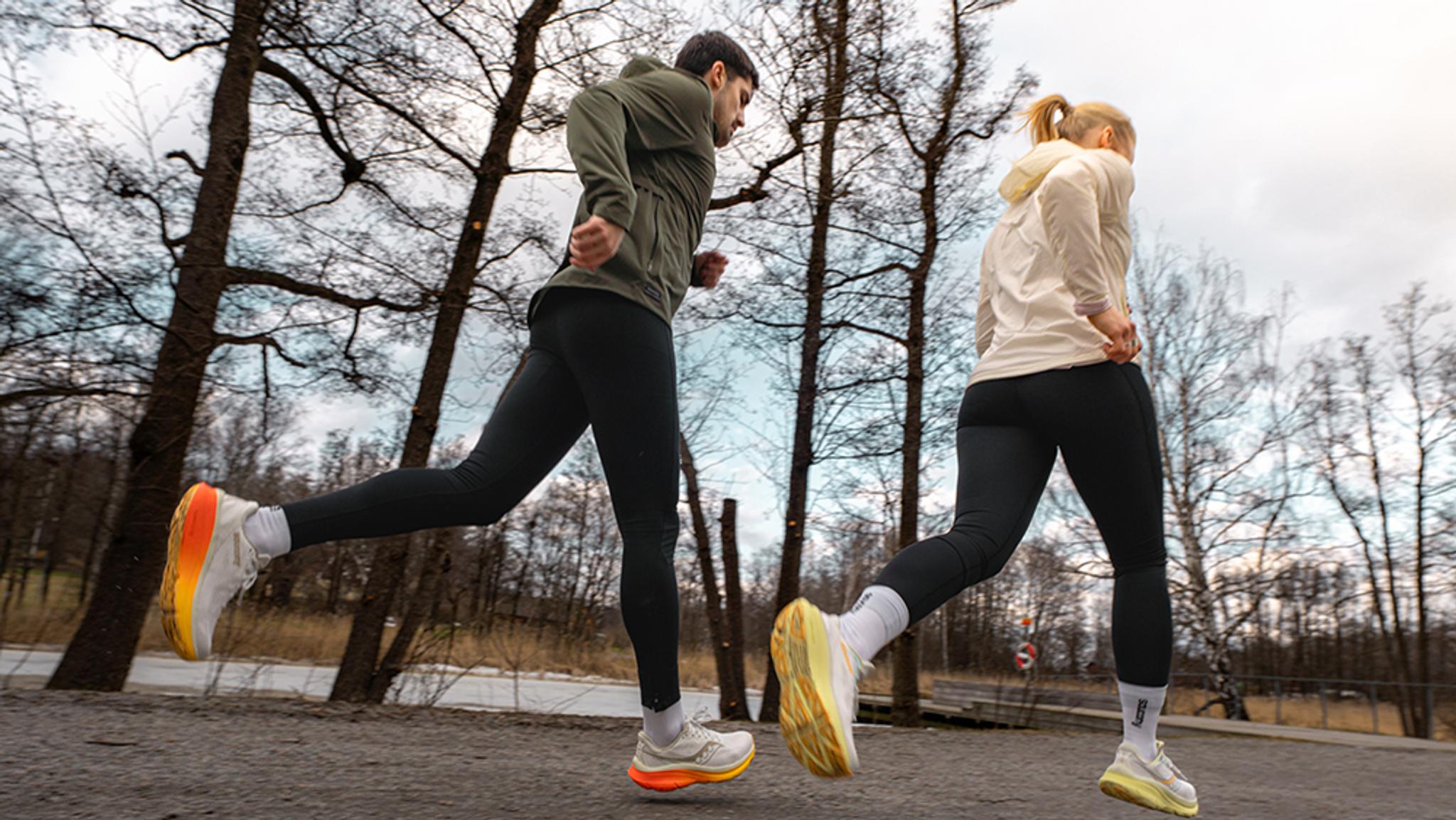 A man and a woman run side-by-side on an outdoor trail, wearing activewear and brightly-soled running shoes, with bare trees in the background.