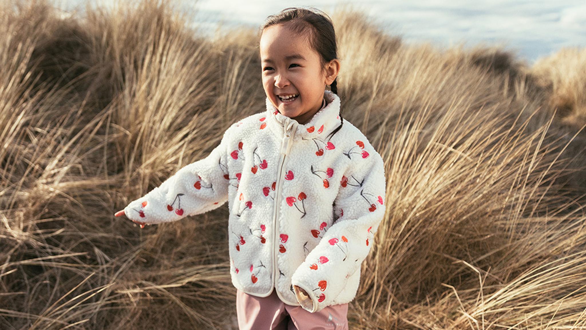 A young girl in a cherry-patterned fuzzy jacket smiles broadly in front of tall dry grass.