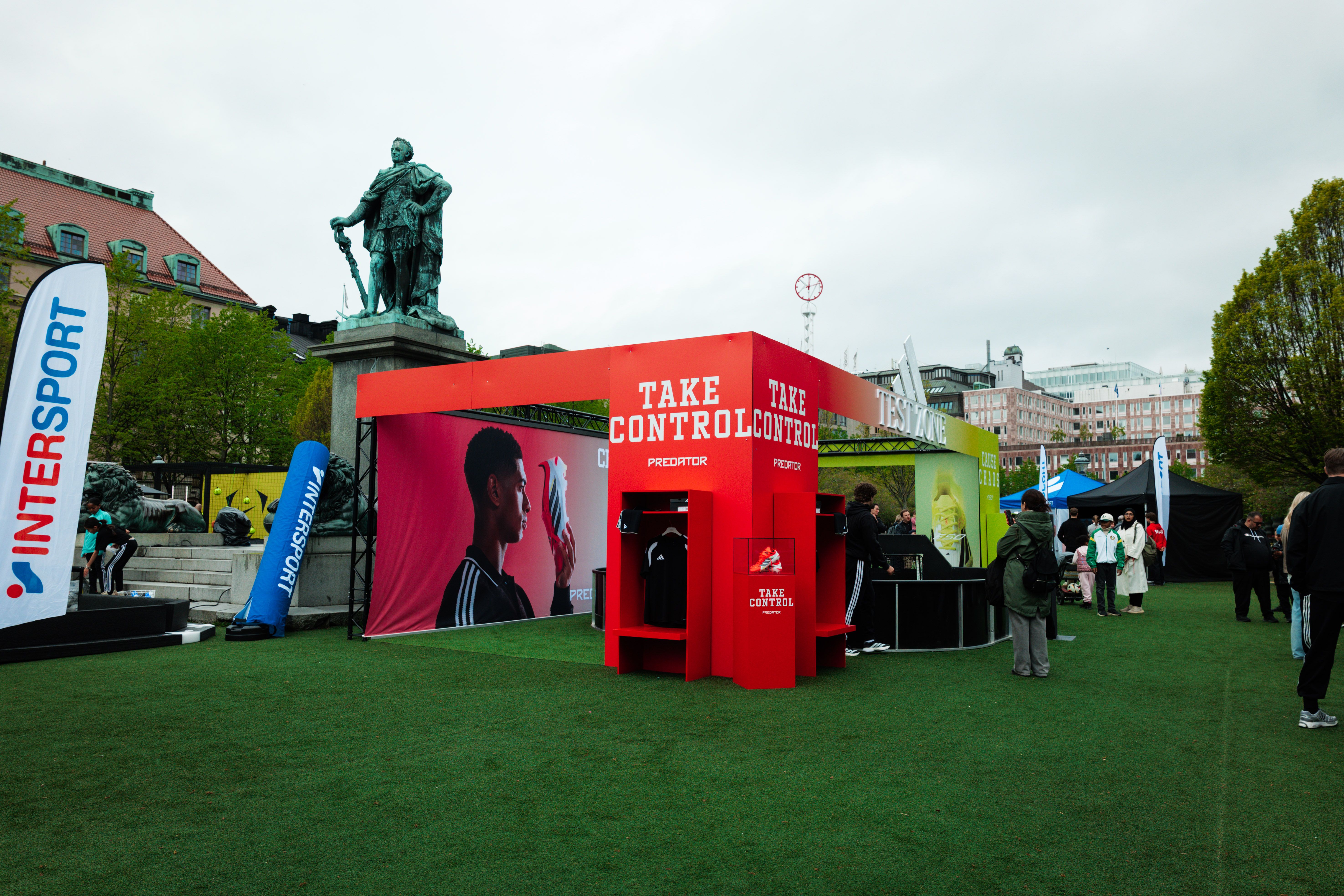 An outdoor event on artificial turf featuring a red 'TAKE CONTROL PREDATOR' display, Intersport branding, and a large statue in the background.