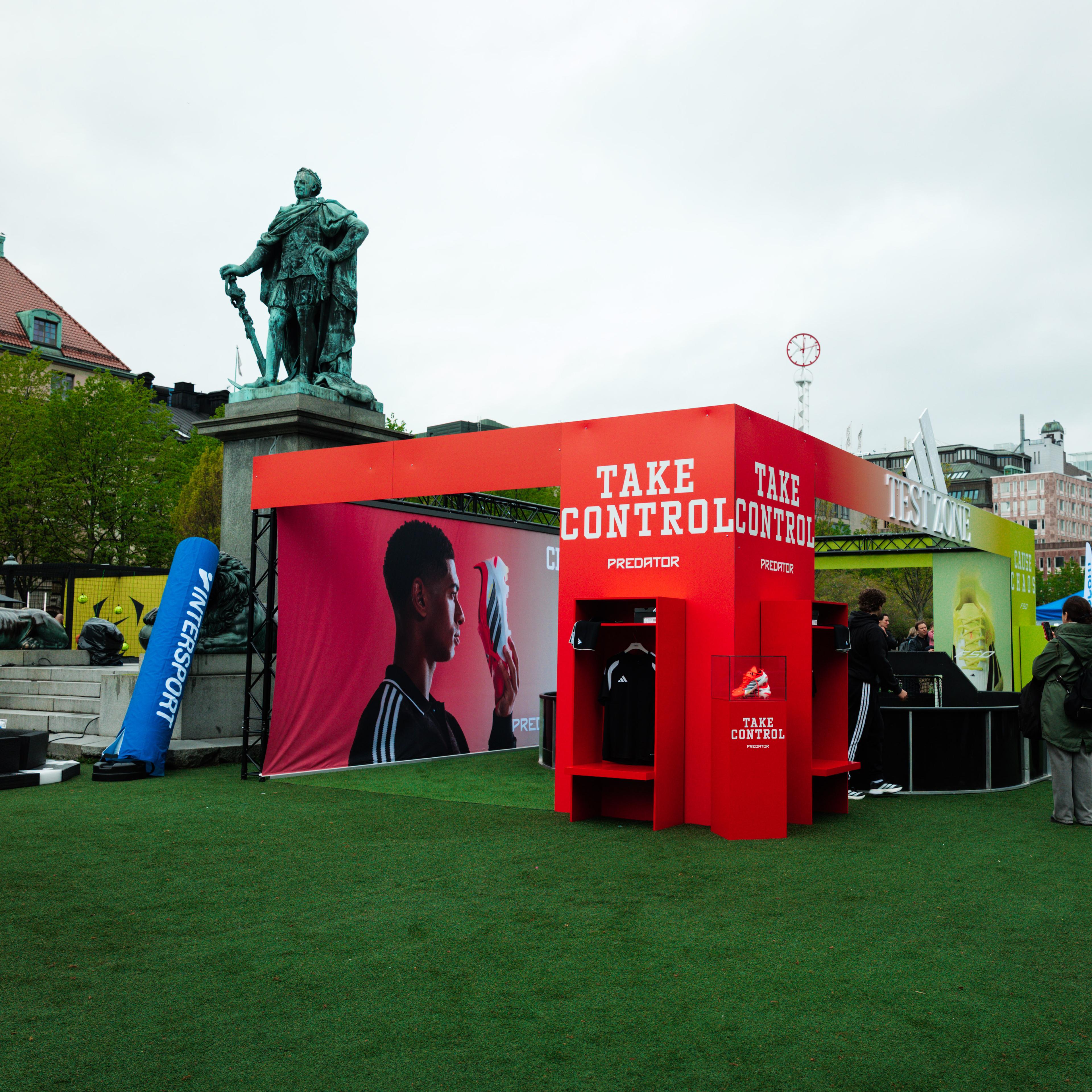 An outdoor event on artificial turf featuring a red 'TAKE CONTROL PREDATOR' display, Intersport branding, and a large statue in the background.