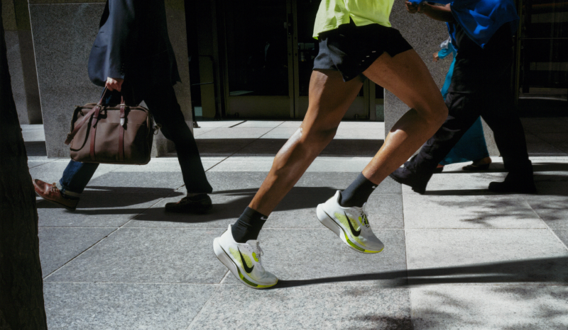 A person in a bright yellow shirt, black shorts, and white running shoes with neon accents runs on a city sidewalk, passing other pedestrians.