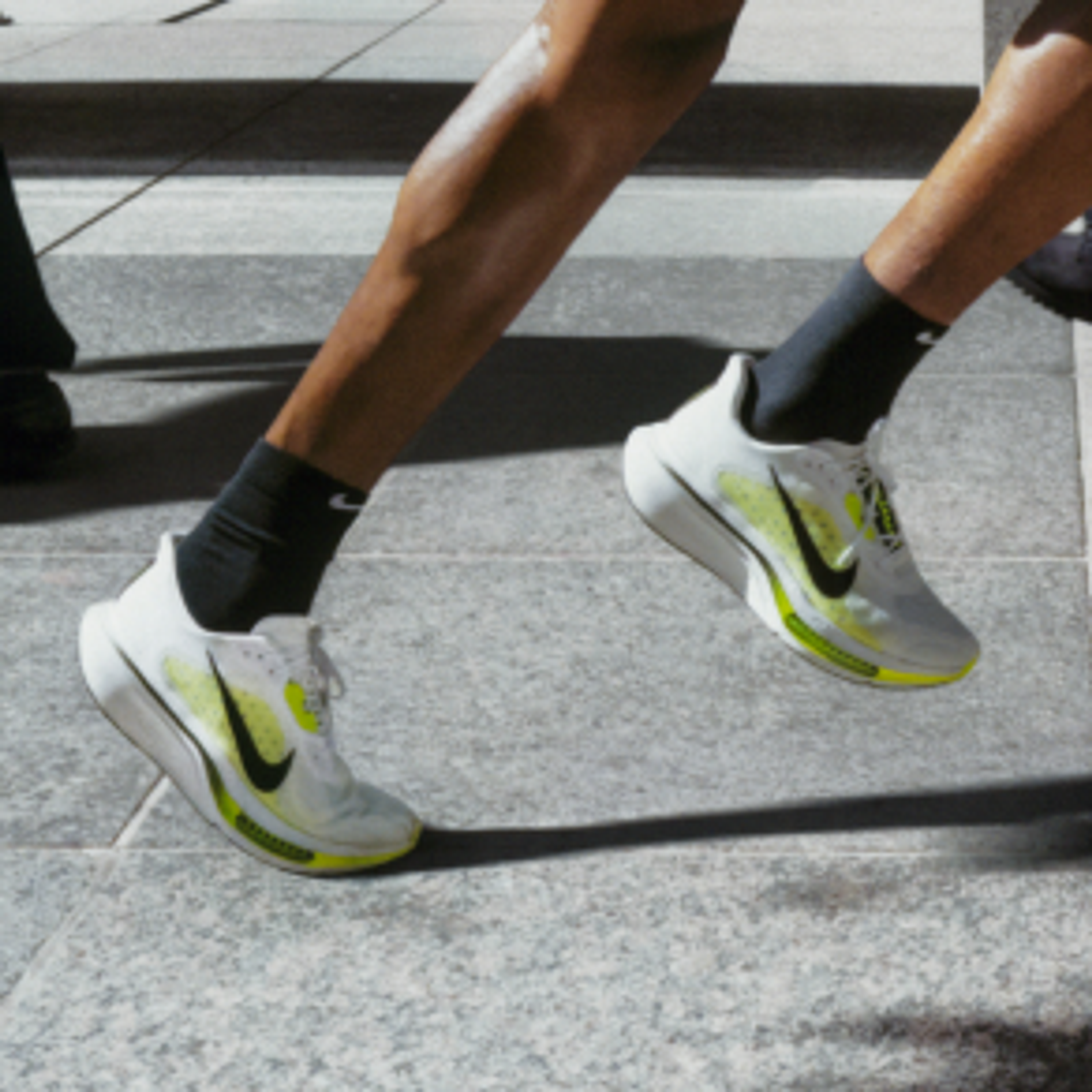 A person in a bright yellow shirt, black shorts, and white running shoes with neon accents runs on a city sidewalk, passing other pedestrians.
