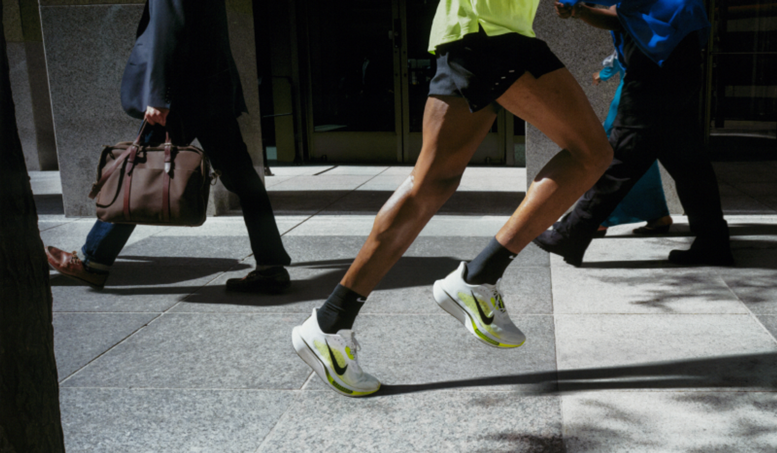 A person in running shorts and bright shoes sprints across a busy sidewalk, past blurred pedestrians.