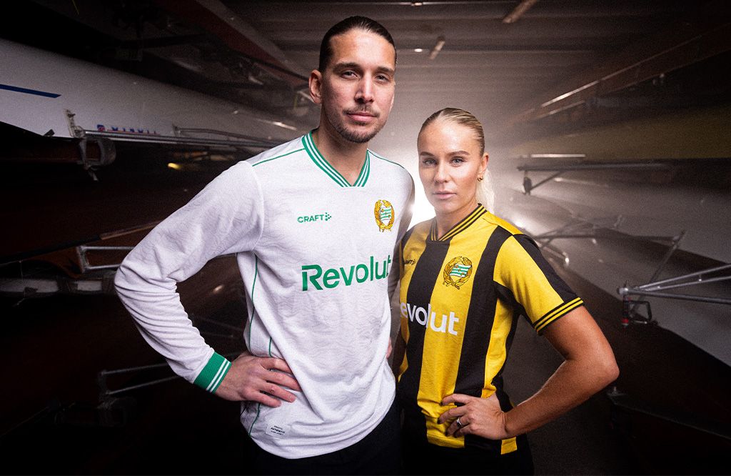 A man in a white and green jersey and a woman in a black and yellow striped jersey stand in a boathouse.