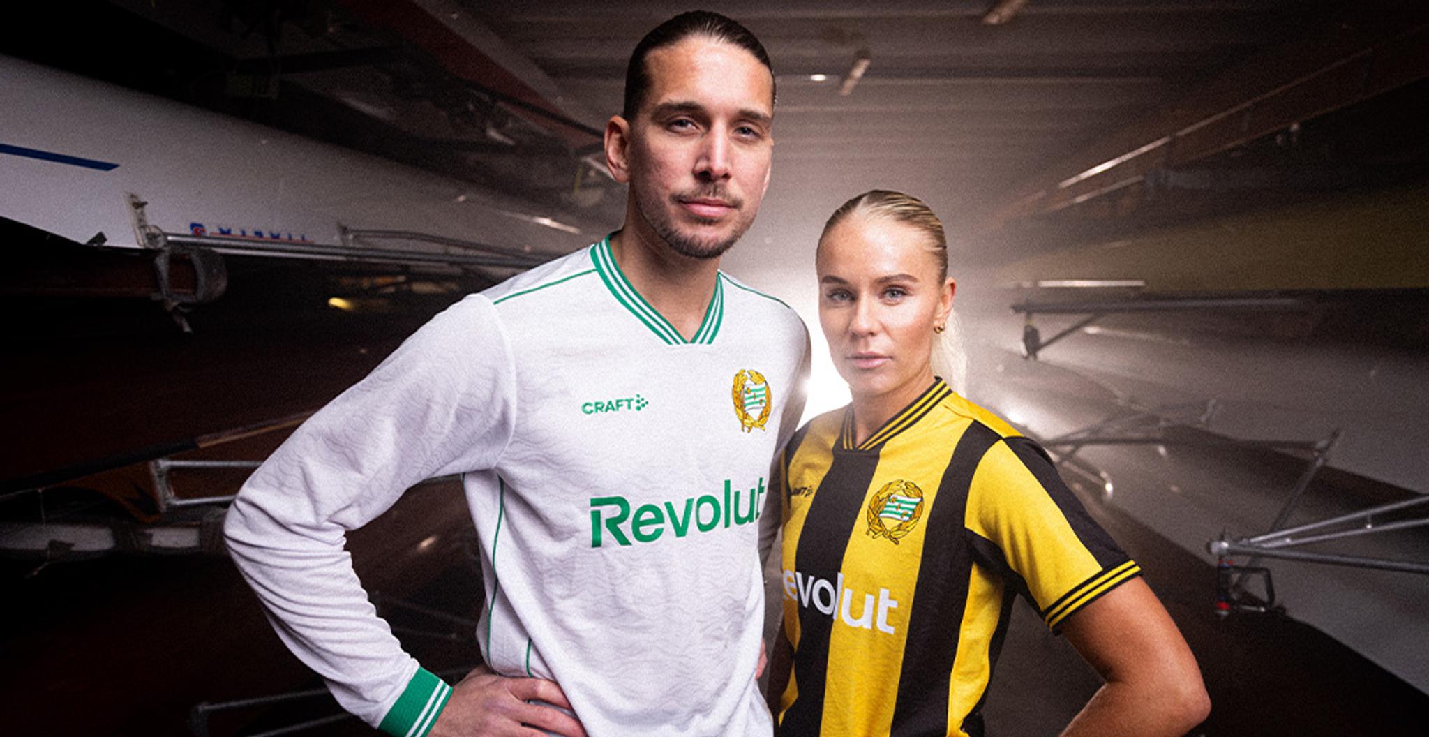 A man in a white and green jersey and a woman in a black and yellow striped jersey stand in a boathouse.