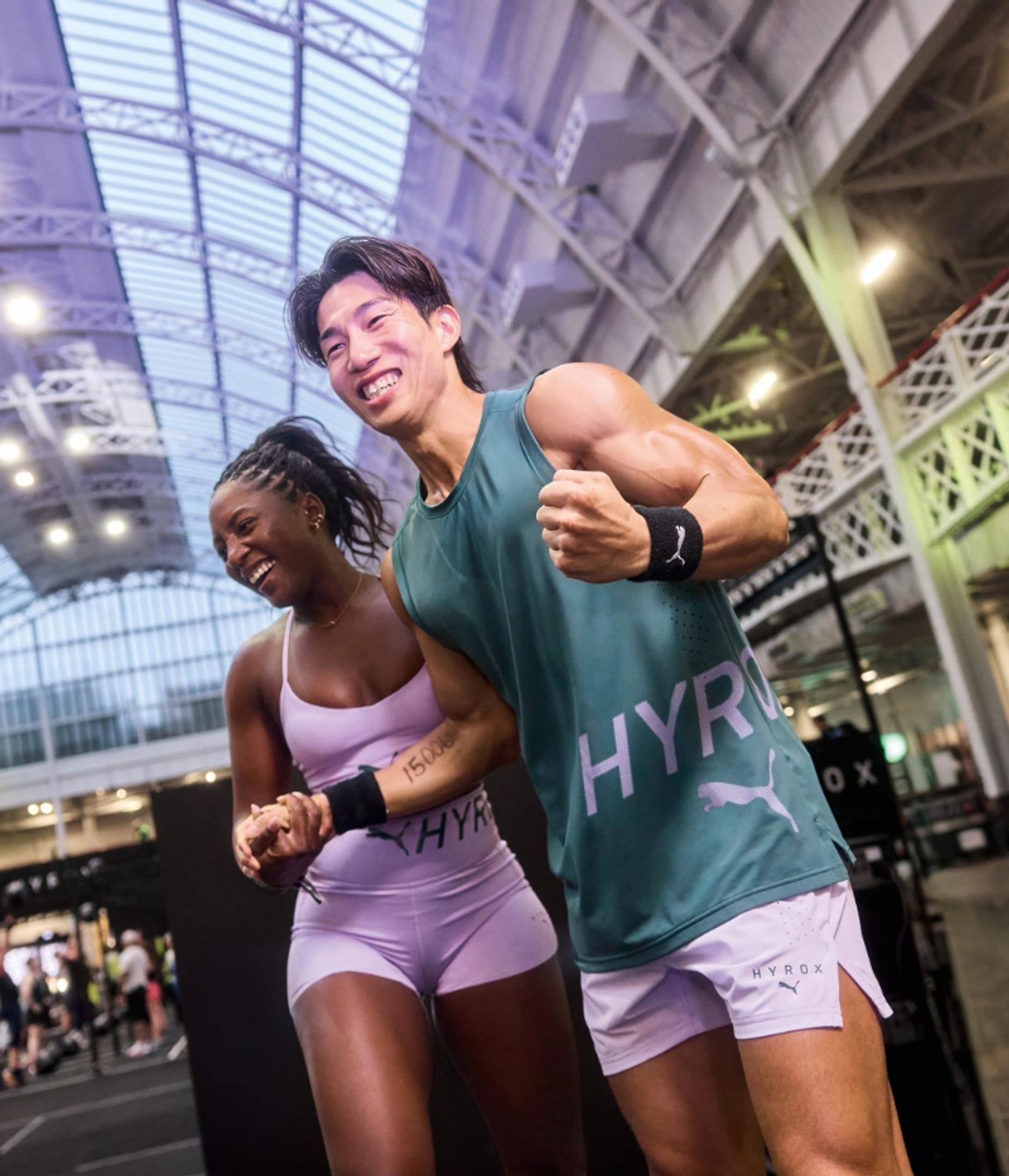 A focused woman in a purple sports bra and shorts pulls down on exercise equipment, with a man blurred in the background.