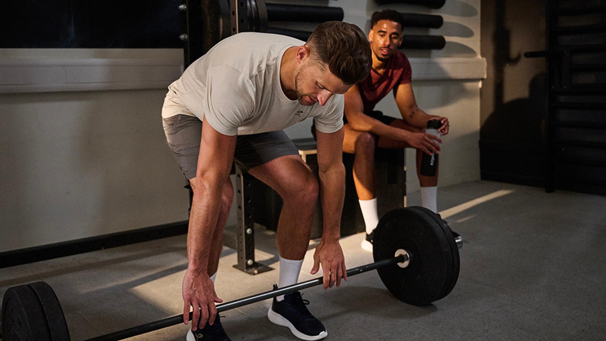 A man prepares to lift a barbell while another man watches in a gym.