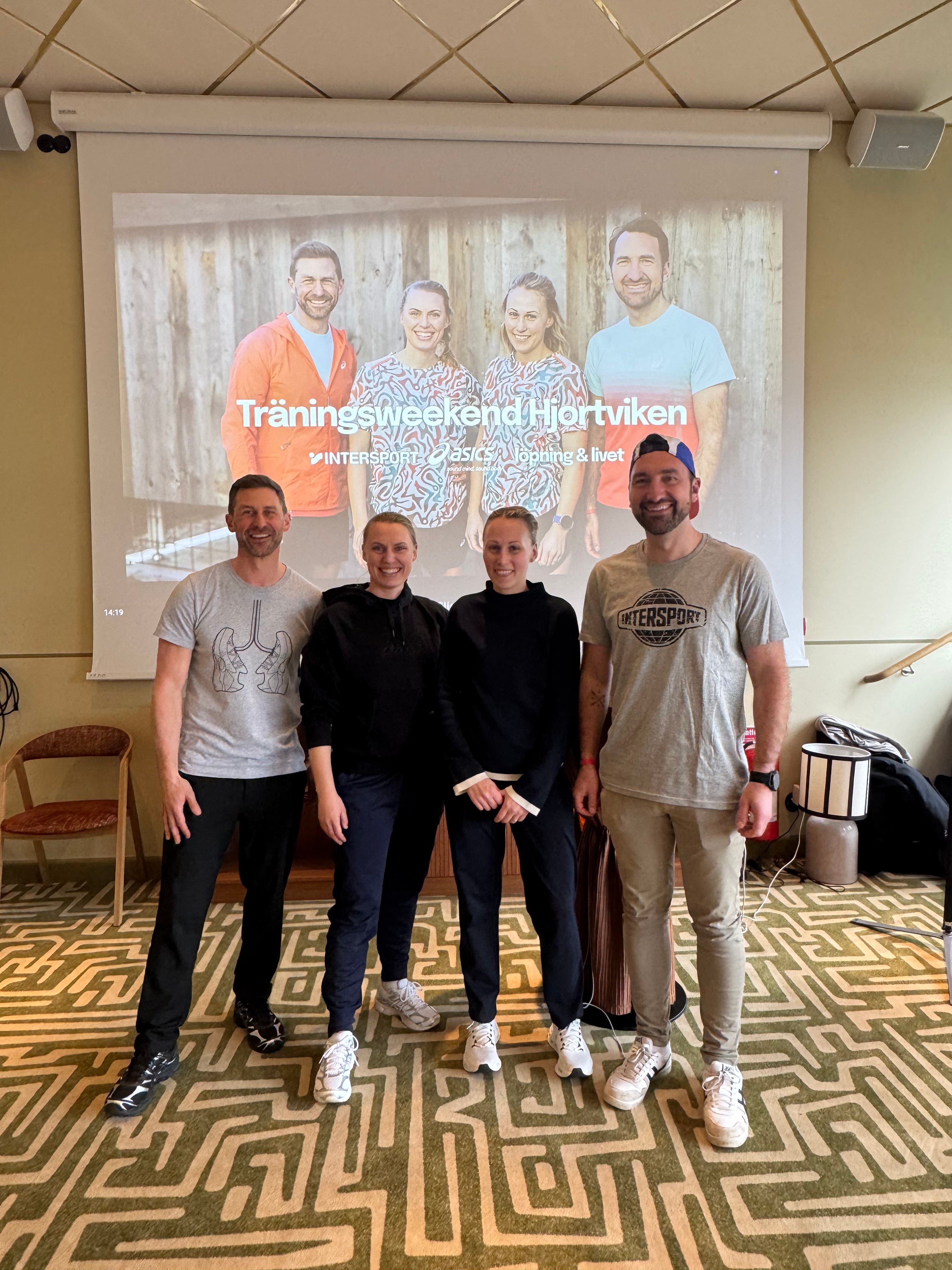 Four smiling people stand in front of a projector screen displaying an event banner for "Träningsweekend Hjortviken" with Intersport and ASICS logos.