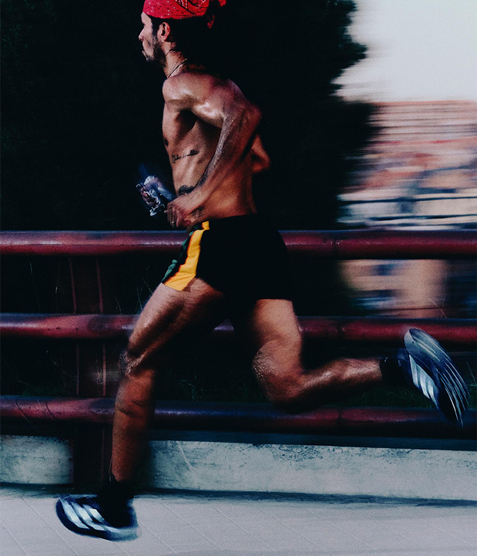 A shirtless man in a red bandana runs quickly, holding a water bottle.