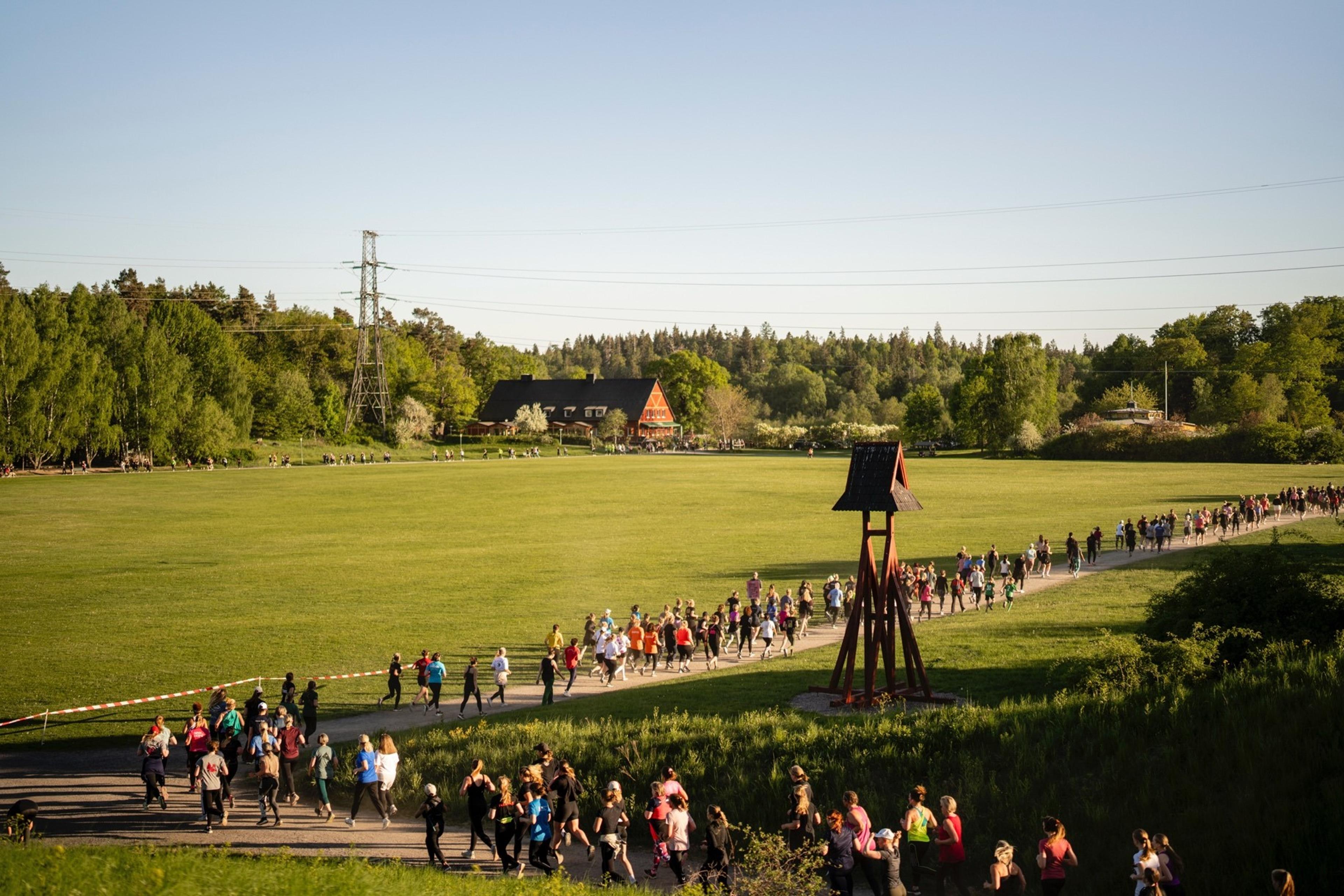 A long line of people walk on a path through a vast green field with a prominent wooden bell tower, surrounded by trees under a clear sky.